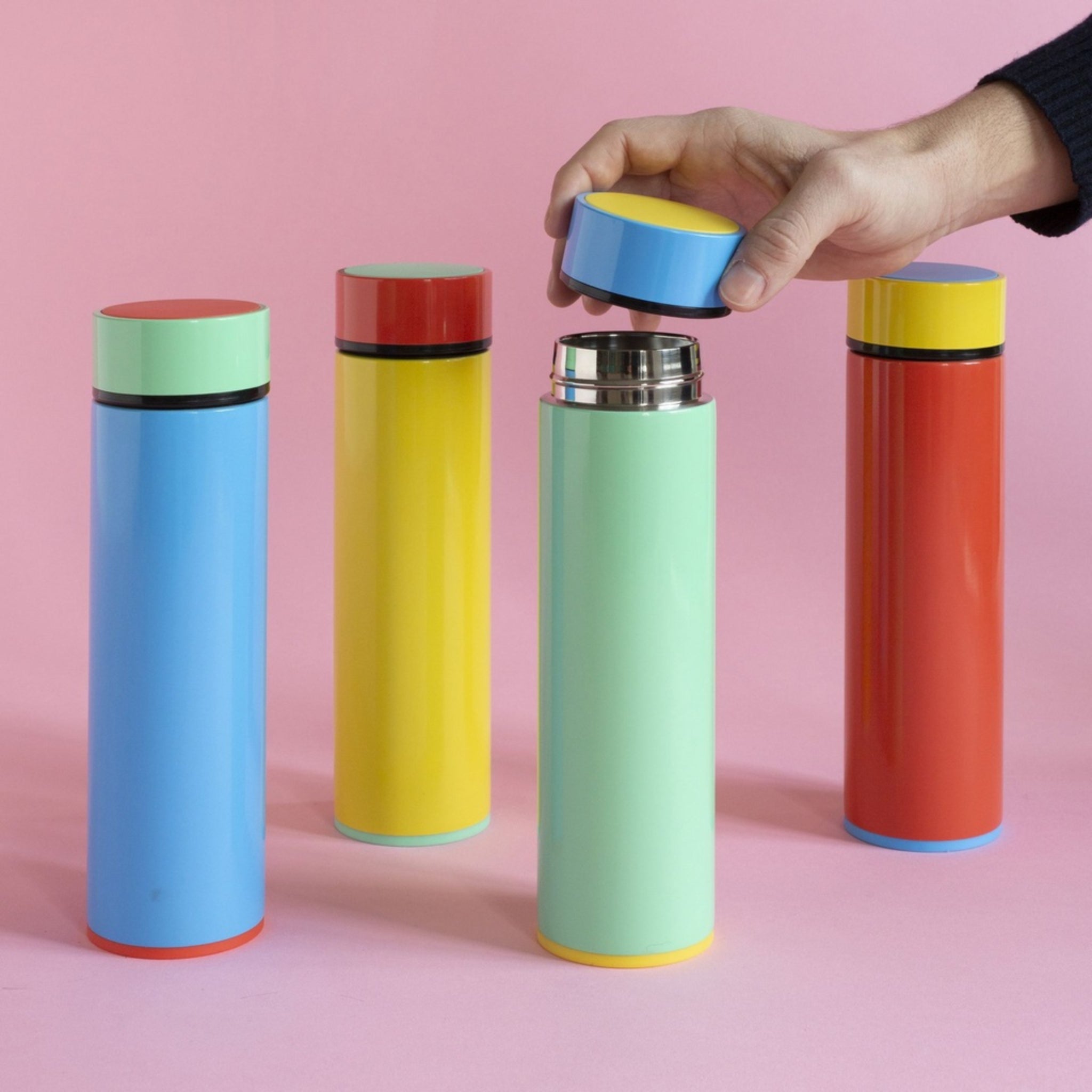 Vier kleurrijke Sowden Thermosflessen staan op een roze ondergrond. Een hand draait het deksel van de blauwe Sowden Thermosfles Blauw 400 ml open, waardoor de metalen binnenkant zichtbaar wordt. De flessen zijn geel, blauw, groen en rood met contrasterende deksels.