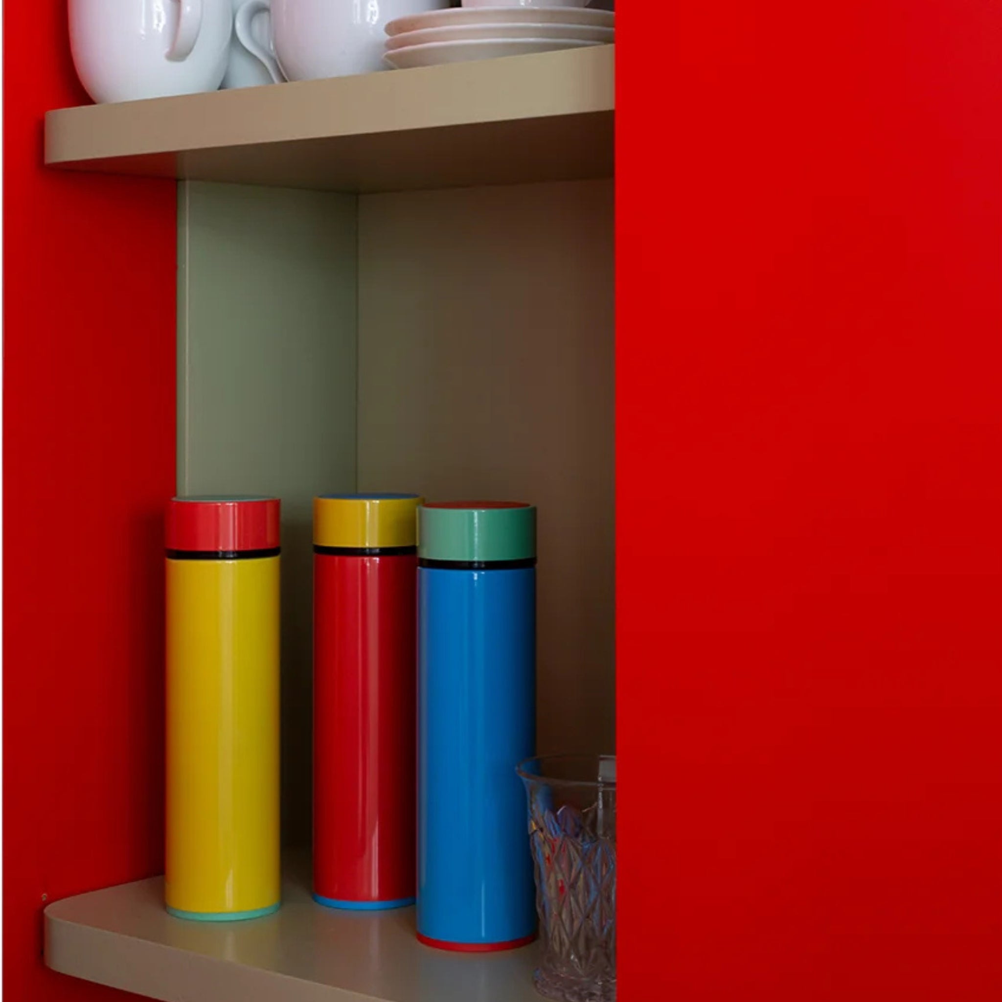 Een Sowden Thermosfles Blauw 400 ml staat op een plank in een kast met rode deuren, ontworpen door George Sowden. Het staat onder witte kopjes en borden, met een glas ernaast.