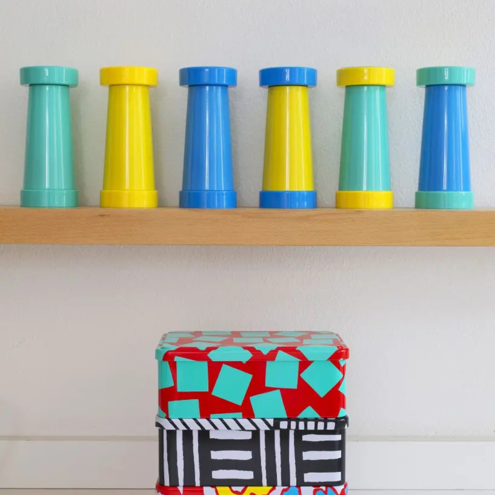 Zes zout- en pepermolens van Sowden Zout-en Pepermolen Blauw, geïnspireerd door de Memphis Design Movement en George Sowden, staan op een houten plank boven twee gedessineerde dozen - de ene rood met blauwe vormen, de andere met zwarte en witte strepen.