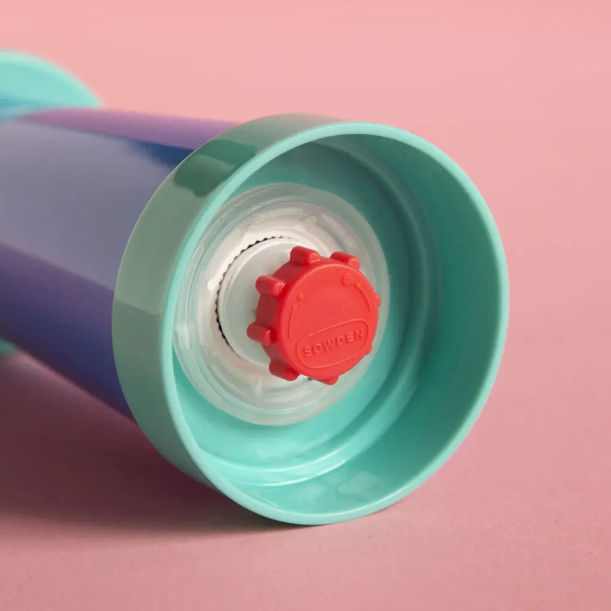 Een close-up van de Sowden Zout-en Pepermolen Veelkleurig van Sowden, een blauw en turquoise cilinder met een rode dop met tekstopdruk en een wit-turquoise rand, tegen een roze achtergrond. Ontworpen door George Sowden.