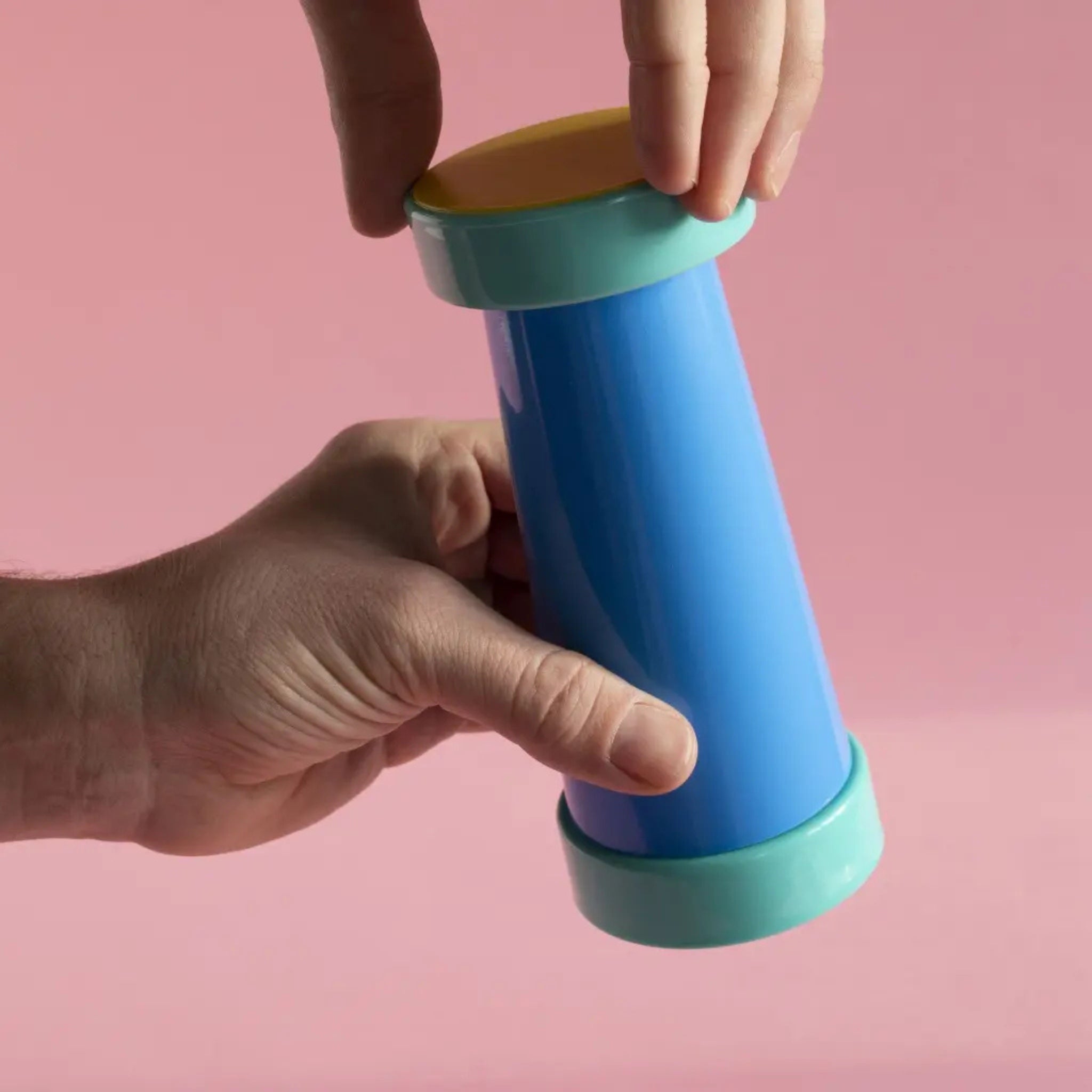 Twee handen houden een Sowden Zout-en Pepermolen Veelkleurig vast tegen een roze achtergrond. De een pakt de zijkant vast, de ander raakt de gele bovenkant aan, wat het speelse ontwerp en keramieken draaimechanisme van Sowden benadrukt.