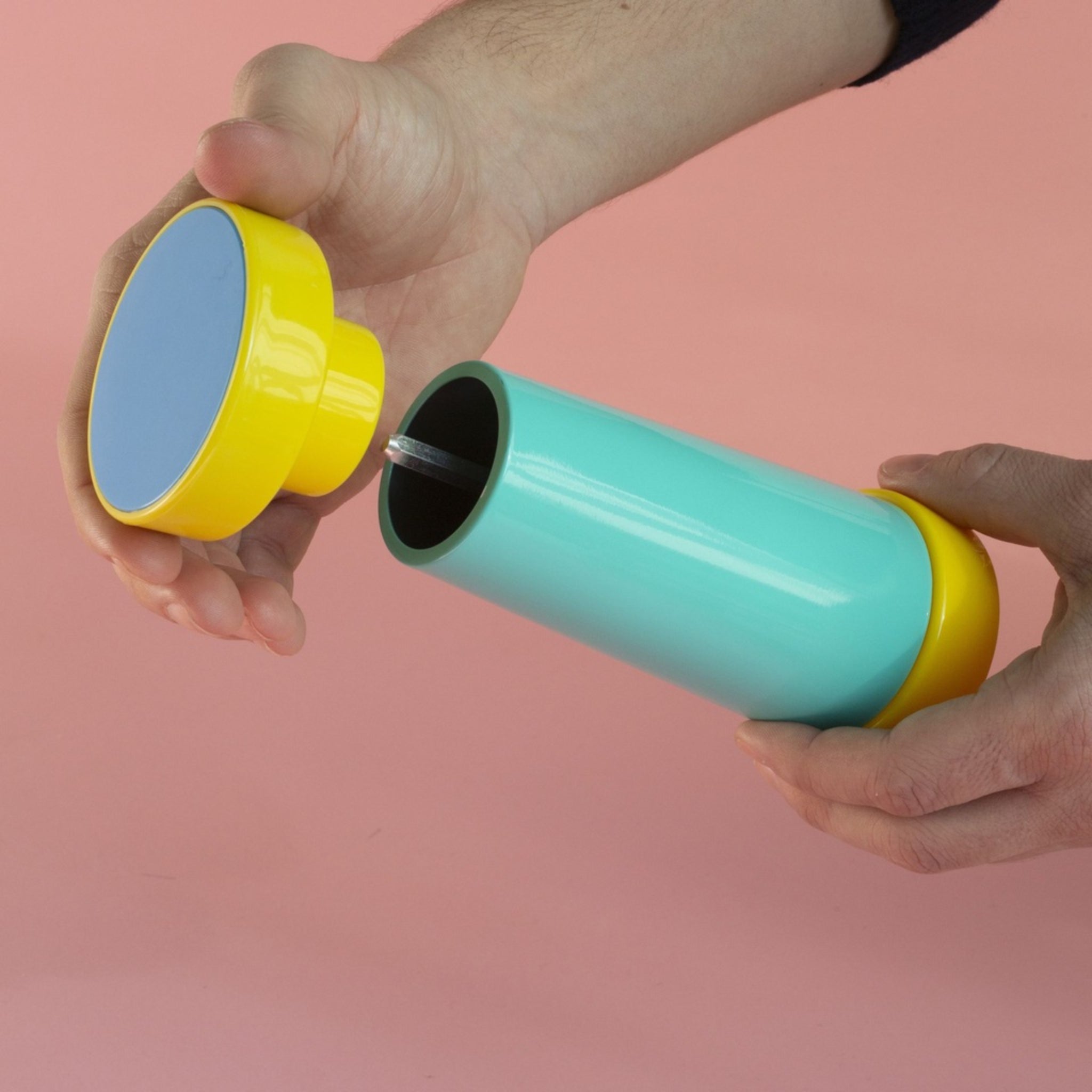 Twee handen houden een cilindervormige Sowden Zout-en Pepermolen Veelkleurig met gele uiteinden vast, één tilt het deksel op om de keramieken draaimechanisme binnenin te laten zien, allemaal tegen een roze achtergrond. Merk: Sowden.