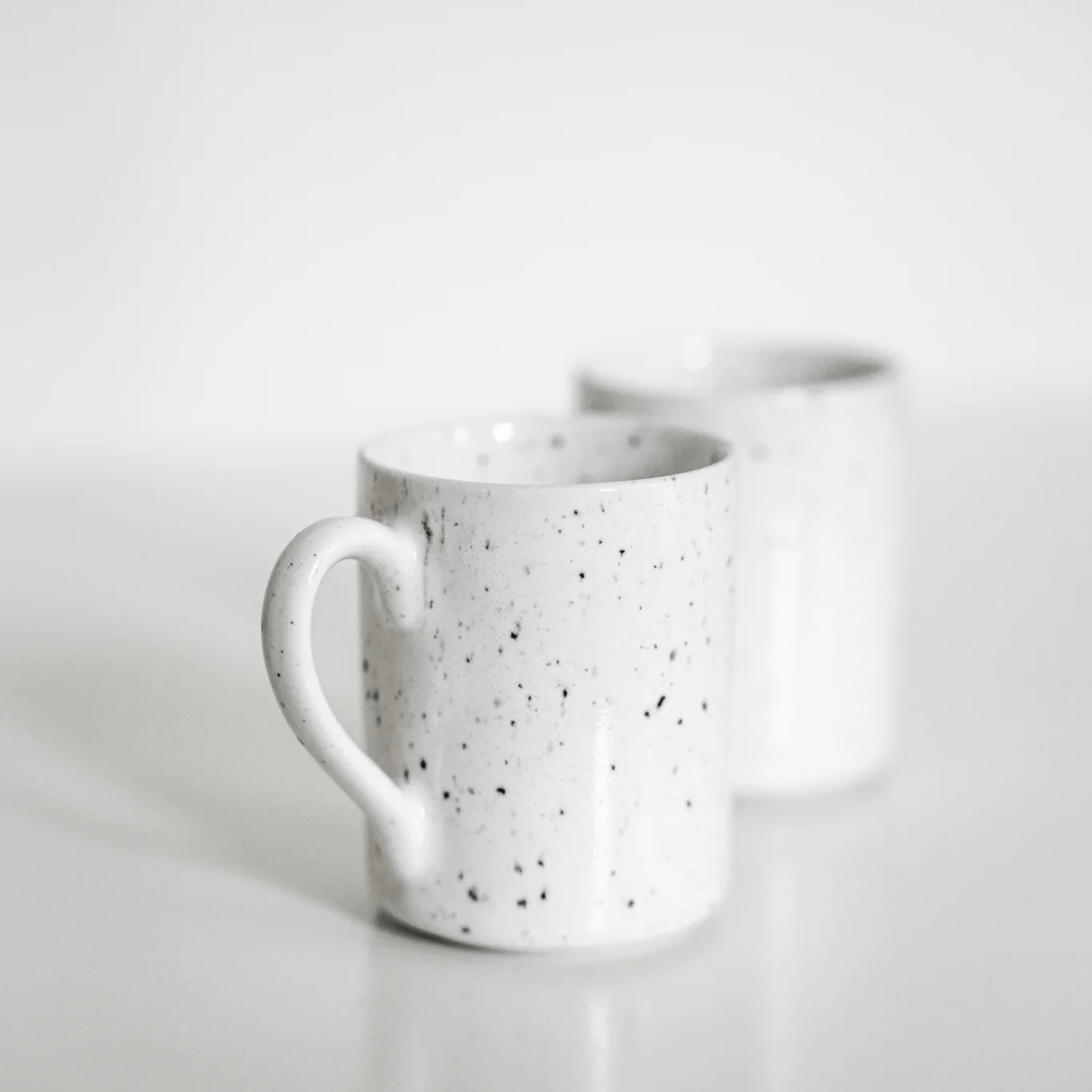 Twee Stock Antwerpen Espresso kop Wit Spikkel mokken van Stock Antwerpen zijn geplaatst op een wit vlak. Het handvat van de mok op de voorgrond is duidelijk zichtbaar, terwijl de andere mok een beetje wazig is achteraan in dit minimalistische tafereel.