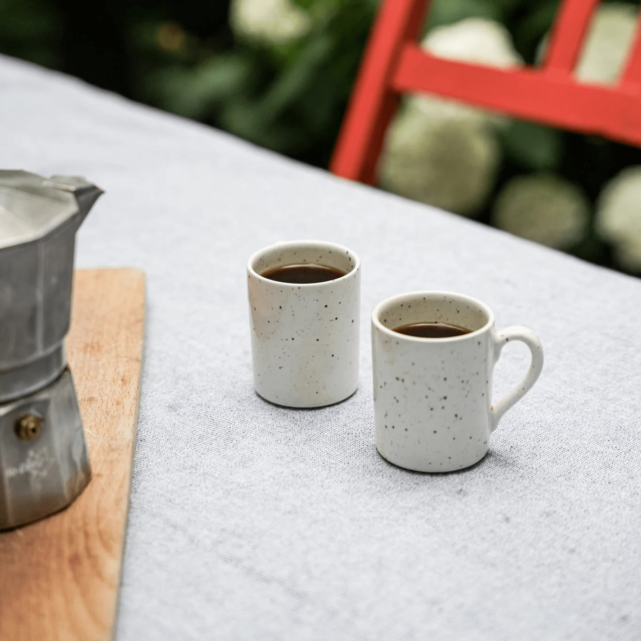 Twee Stock Antwerpen Espresso kop Wit Spikkel, gevuld met koffie, rusten op een lichtgrijs tafelkleed naast een zilveren mokapot en houten plank. Een felrode stoel en weelderig groen maken het tafereel compleet met uniek servies op de achtergrond.
