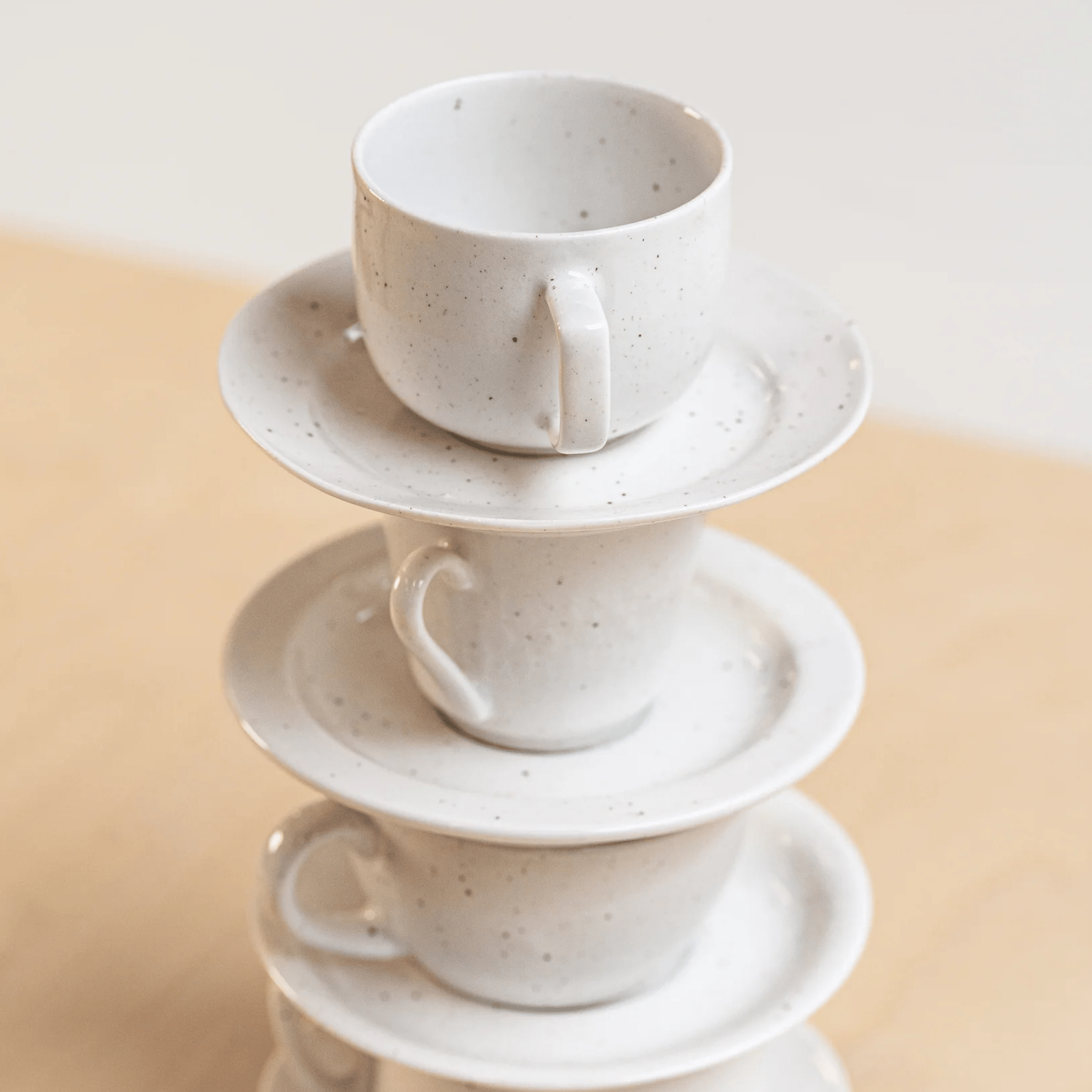 Vier witte Stock Antwerpen Koffiekop + Schotel Wit Spikkel zijn gestapeld op een lichte houten ondergrond. Dit servies van Stock Antwerpen brengt stijl aan elke tafel en is ideaal voor liefhebbers van hergebruikt servies.