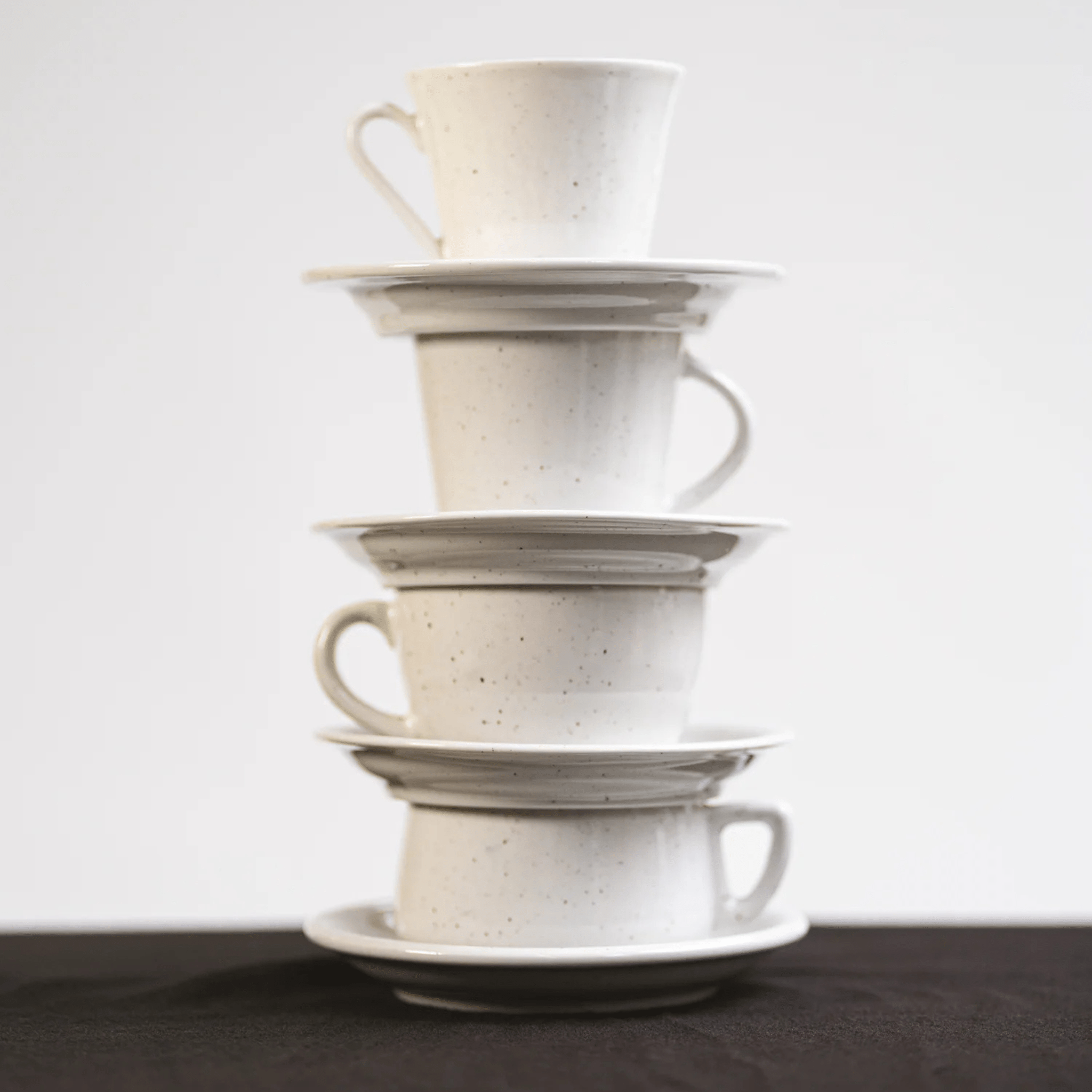 Vier Stock Antwerpen Koffiekop + Schotel Wit Spikkel kopjes zijn gestapeld met schoteltjes en vormen een hoge, licht overhellende toren op een donker oppervlak. Dit benadrukt het unieke ontwerp van Stock Antwerpen tegen een effen witte achtergrond.