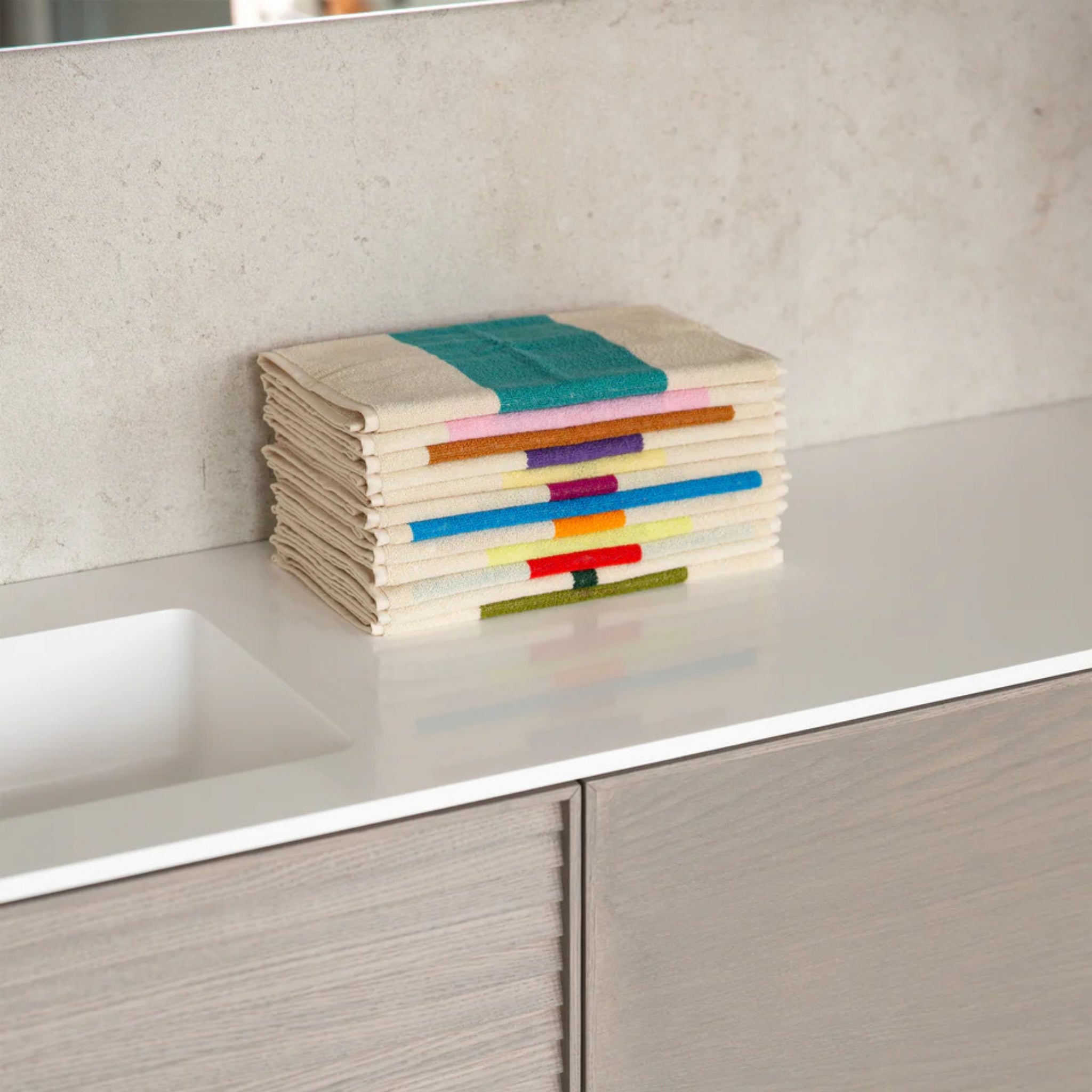 Een netjes opgevouwen Suite 702 Gastendoekjes Set C 30/50 (3 st.) van Suite 702, gemaakt van biologisch katoen met op Karel Martens geïnspireerde kleurrijke strepen, ligt op een wit aanrecht naast een wastafel met lichtgrijze stenen en houten kastjes.