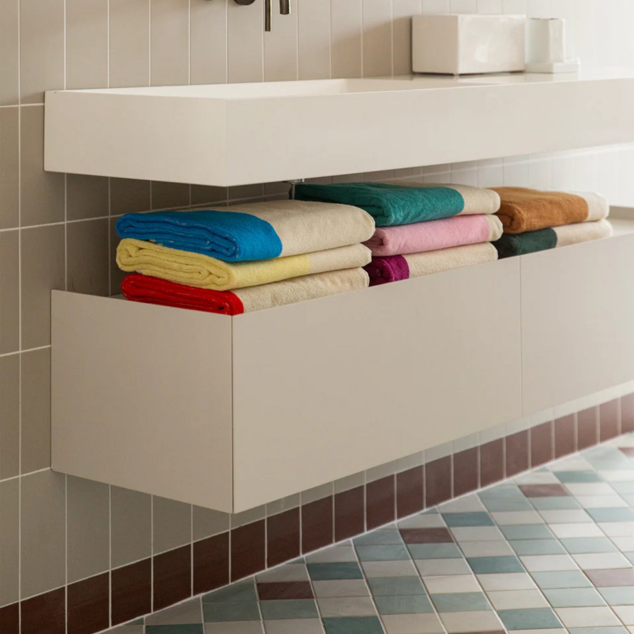 Een moderne badkamer met een zwevend wit badmeubel en een open schap netjes gestapeld met Suite 702 Badhanddoek Rood Turquoise 70/140 handdoeken; de vloer met blauw, wit en bruin patroon weerspiegelt de grafische stijl van Karel Martens.