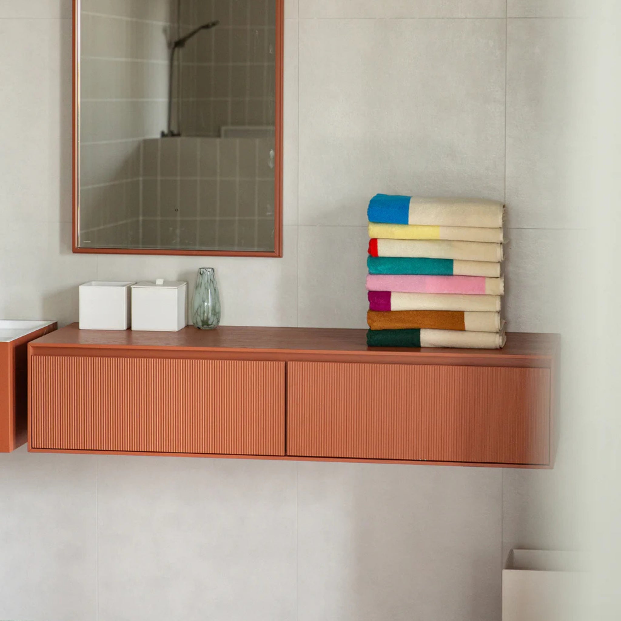 Een moderne badkamer met een zwevend houten badmeubel, een spiegel aan de muur, twee witte containers, een glazen fles en een keurig gestapelde stapel Suite 702 Badhanddoek Rood Turquoise 70/140 van Suite 702 op het aanrecht. Lichtgrijze tegels bedekken de muren.