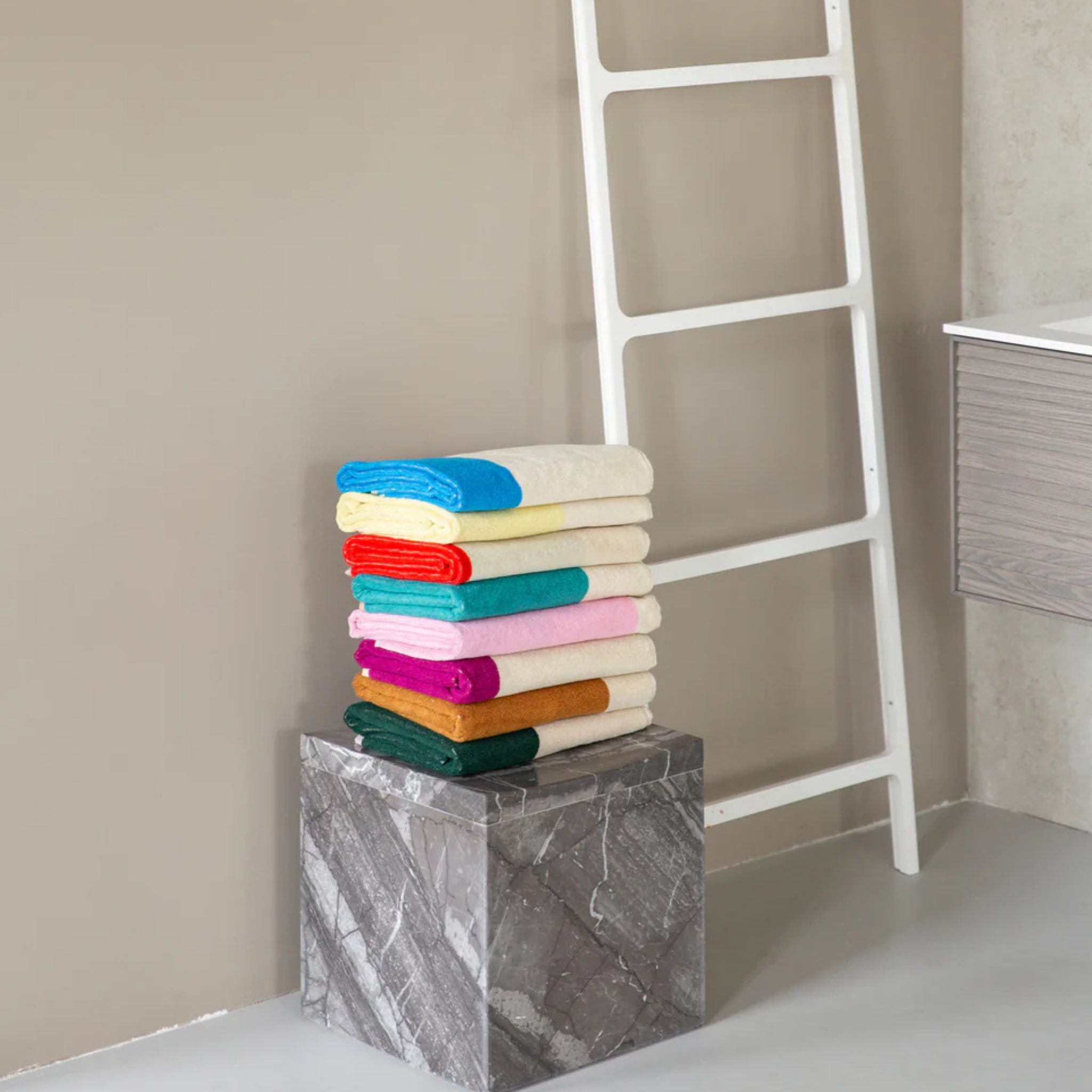 De Suite 702 Badhanddoek Rood Turquoise 70/140 van Suite 702, gemaakt van biologisch katoen, staat op een grijs marmeren sokkel bij een witte ladder en lichtgrijs badmeubel, met een beige muur op de achtergrond.