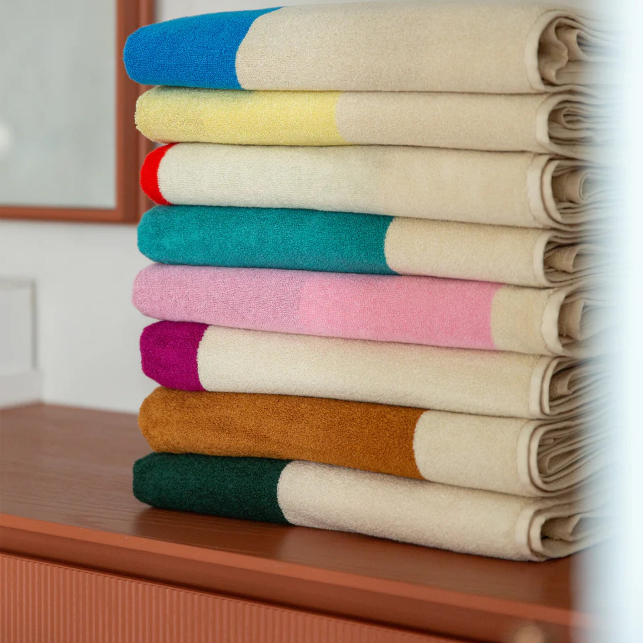 Een netjes gevouwen stapeltje Suite 702 Badhanddoek Aqua Toffee 70/140 van Suite 702, gemaakt van biologisch katoen met crèmekleurige delen en gekleurde uiteinden, gerangschikt op een houten oppervlak met een onscherpe achtergrond.