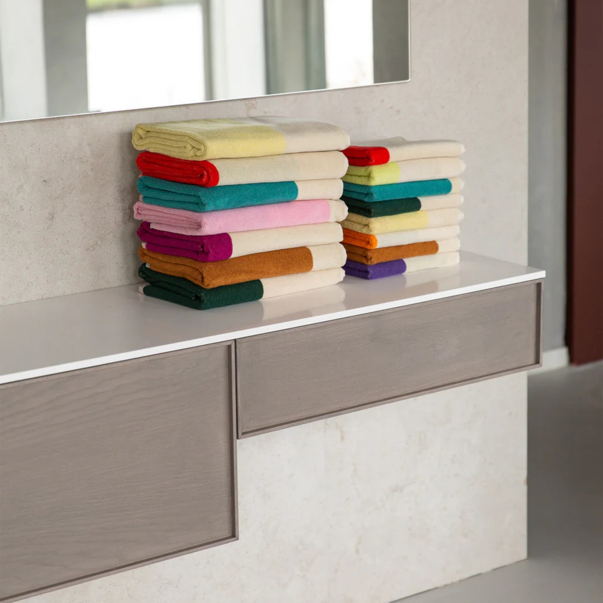 Twee nette stapels Suite 702 Badhanddoek Rood Turquoise 70/140 gemaakt van biologisch katoen liggen op een modern wit aanrecht in een badkamer, met een grote spiegel en lichthouten kasten op de achtergrond.