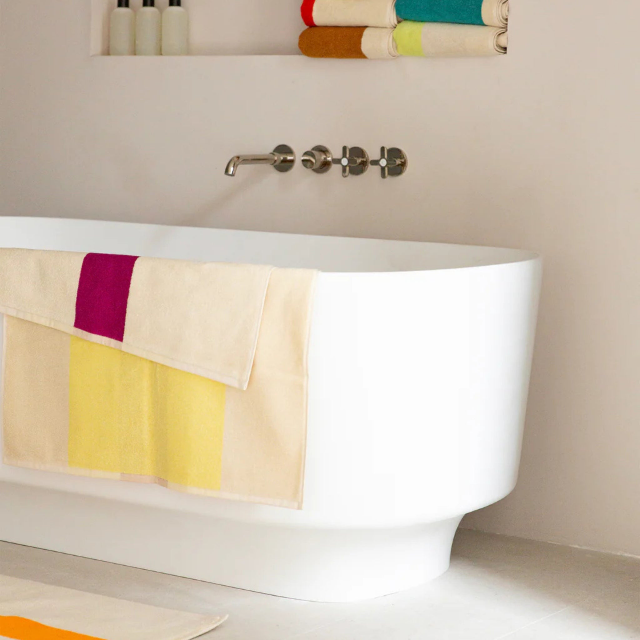 Een modern wit bad heeft een Suite 702 Badhanddoek Geel Fuchsia 70/140 gedrapeerd over de zijkant. Op een plank erboven liggen opgerolde handdoeken van biologisch katoen, met flessen netjes in de buurt.