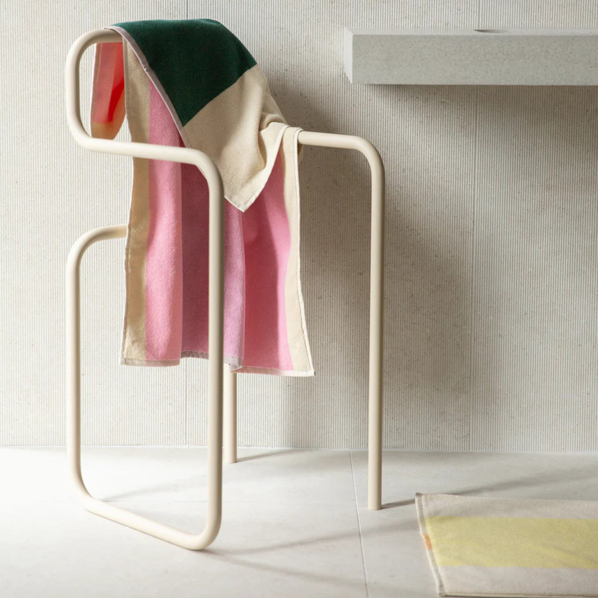 De Suite 702 Badhanddoek Roze Donkergroen 70/140, een handdoek met geometrisch patroon in roze, groen en crème van biologisch katoen, is gedrapeerd over een moderne witte metalen stoel op lichte tegels bij een zwevend aanrecht.
