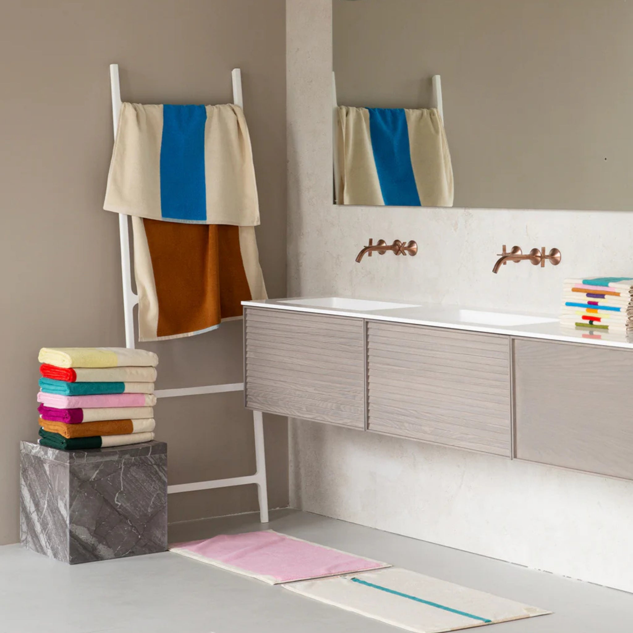 Een moderne badkamer met een dubbele wastafel, wandspiegel en een opgevouwen Suite 702 Badhanddoek Aqua Toffee 70/140 op een marmeren blok, aangevuld met een witte ladder met gedrapeerde handdoeken en een gestreepte badmat op de vloer.