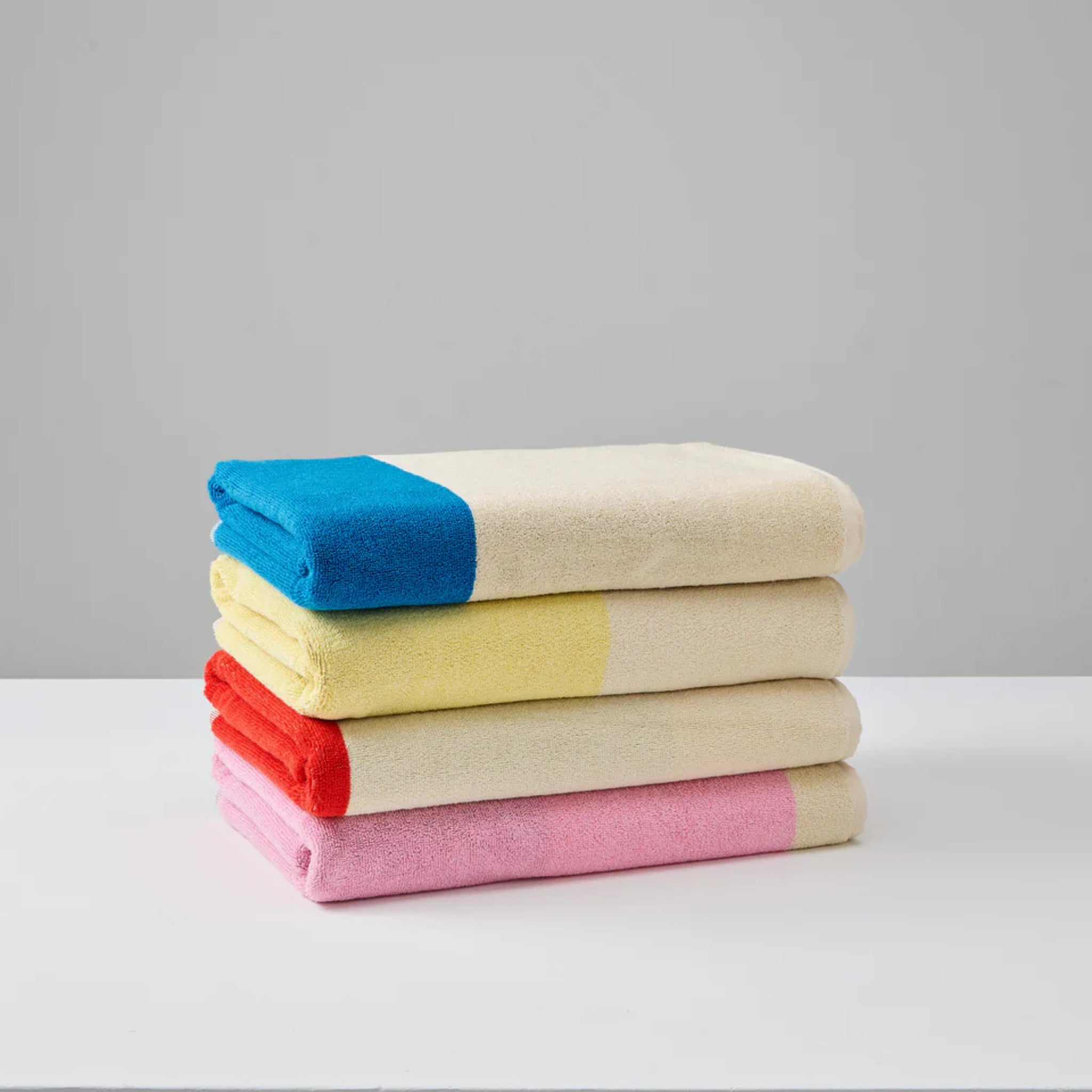 Een stapel van vier Suite 702 Badhanddoeken Rood Turquoise 70/140, gemaakt van biologisch katoen, ligt op een wit oppervlak met grote gekleurde hoeken - blauw, geel, rood en roze - die contrasteren met de lichtbeige stof.