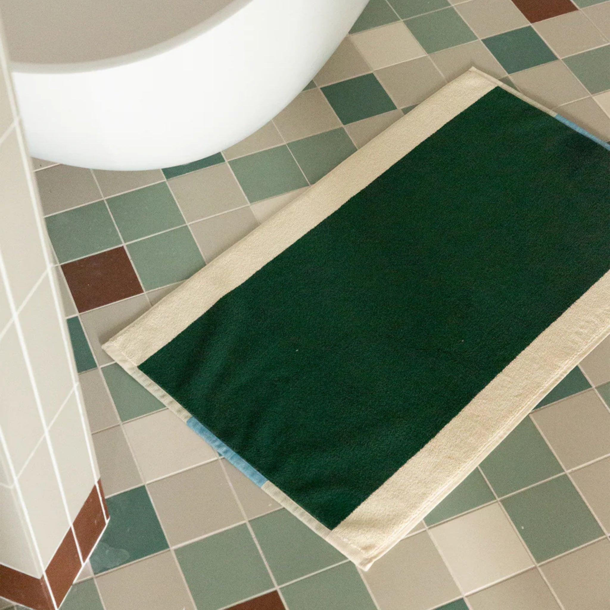 De Suite 702 Badmat Aqua Donkergroen 50/80, een rechthoekige groene badmat van biologisch katoen met crèmekleurige randen, ligt op een betegelde badkamervloer in groene, witte en bruine vierkanten bij een wit bad.