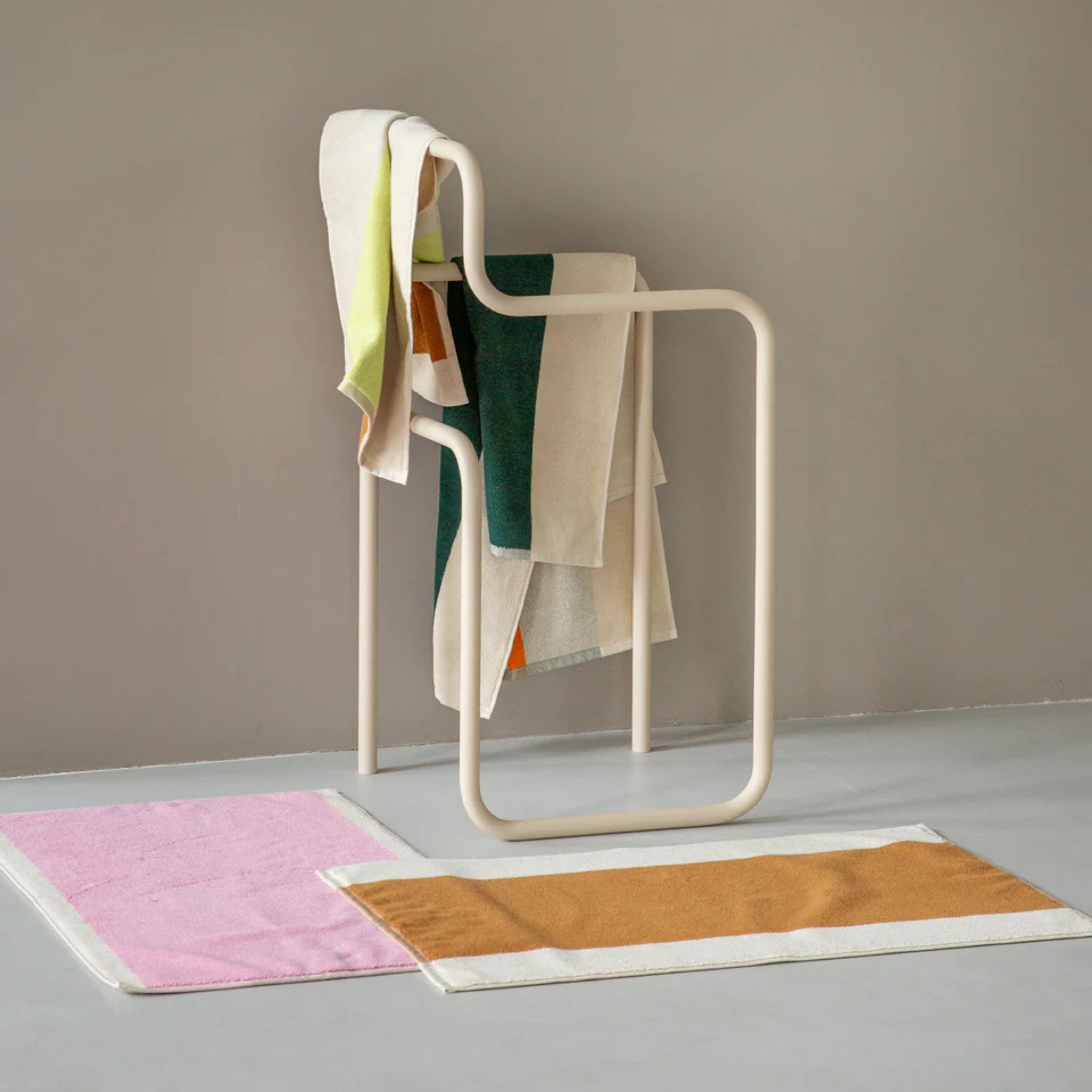 Een minimalistisch crèmekleurig handdoekenrek toont kleurrijke handdoeken. Op de grijze vloer geven twee badmatten van Suite 702 Badmat Toffee Turquoise 50/80 een modern tintje tegen een licht taupekleurige muur, als knipoog naar de levendige stijl van Karel Martens.