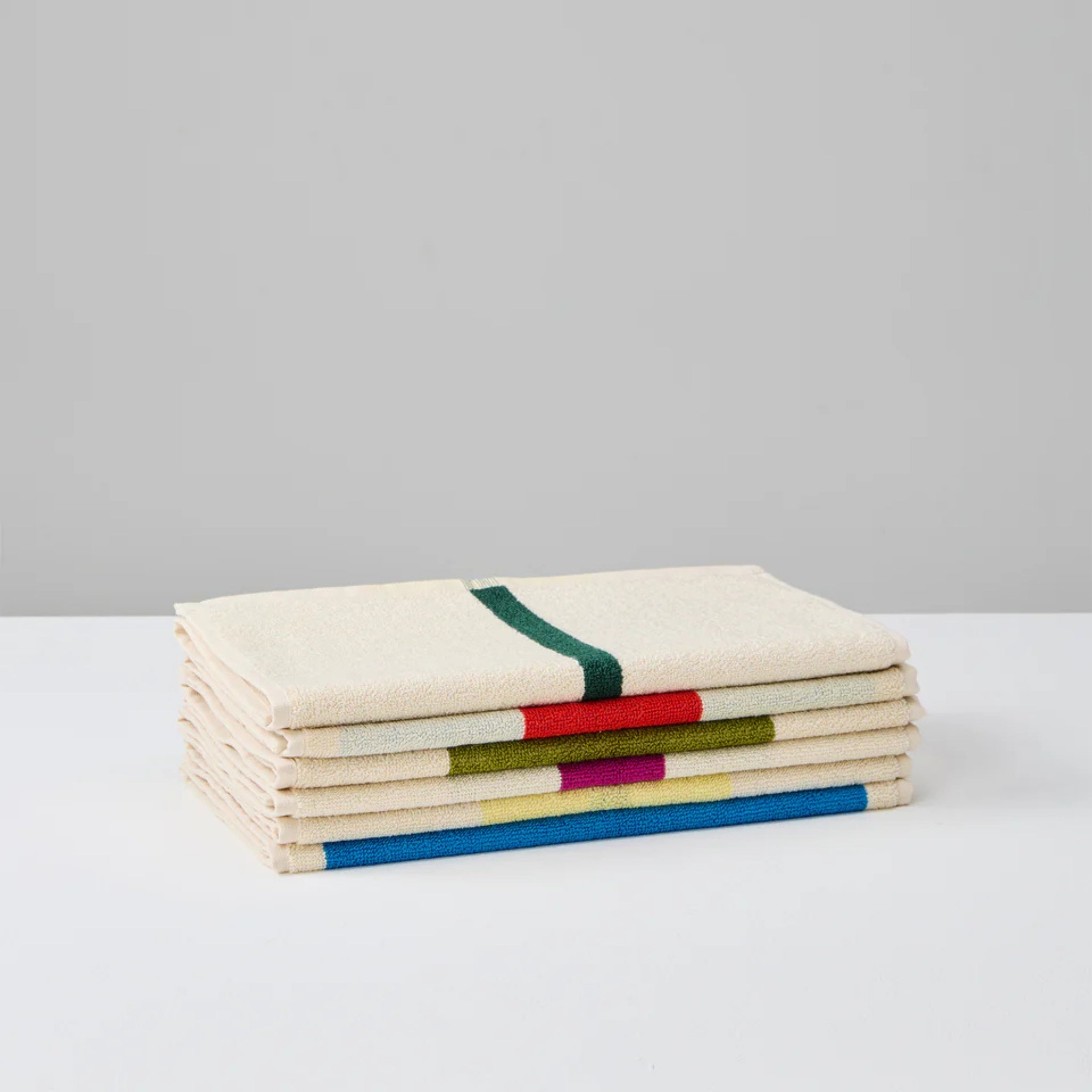 Een set van drie crèmekleurige Suite 702 Gastendoekjes (Set D 30/50) gemaakt van biologisch katoen, elk met minimale kleurrijke geometrische lijnen geïnspireerd door Karel Martens, weergegeven op een wit oppervlak en een lichtgrijze achtergrond.