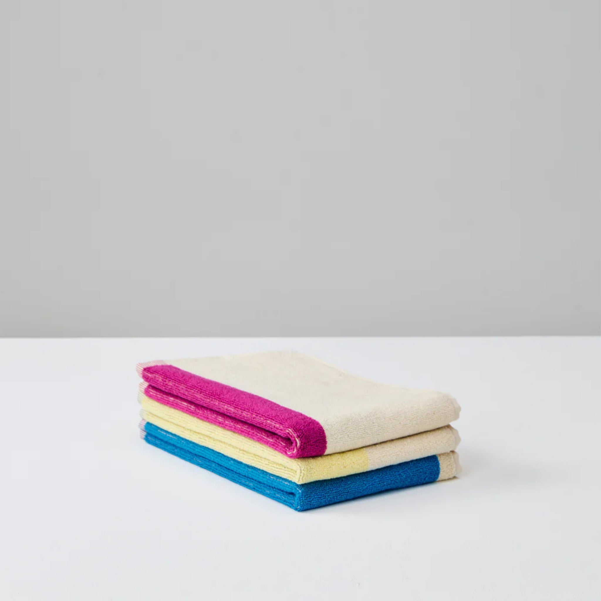 Een netjes gevouwen Suite 702 Gastendoekjes Set D 30/50 (3 st.), gemaakt van biologisch katoen, rust op een wit oppervlak. Elke handdoek heeft een felgekleurde rand - roze, geel en blauw - tegen een effen lichtgrijze achtergrond.