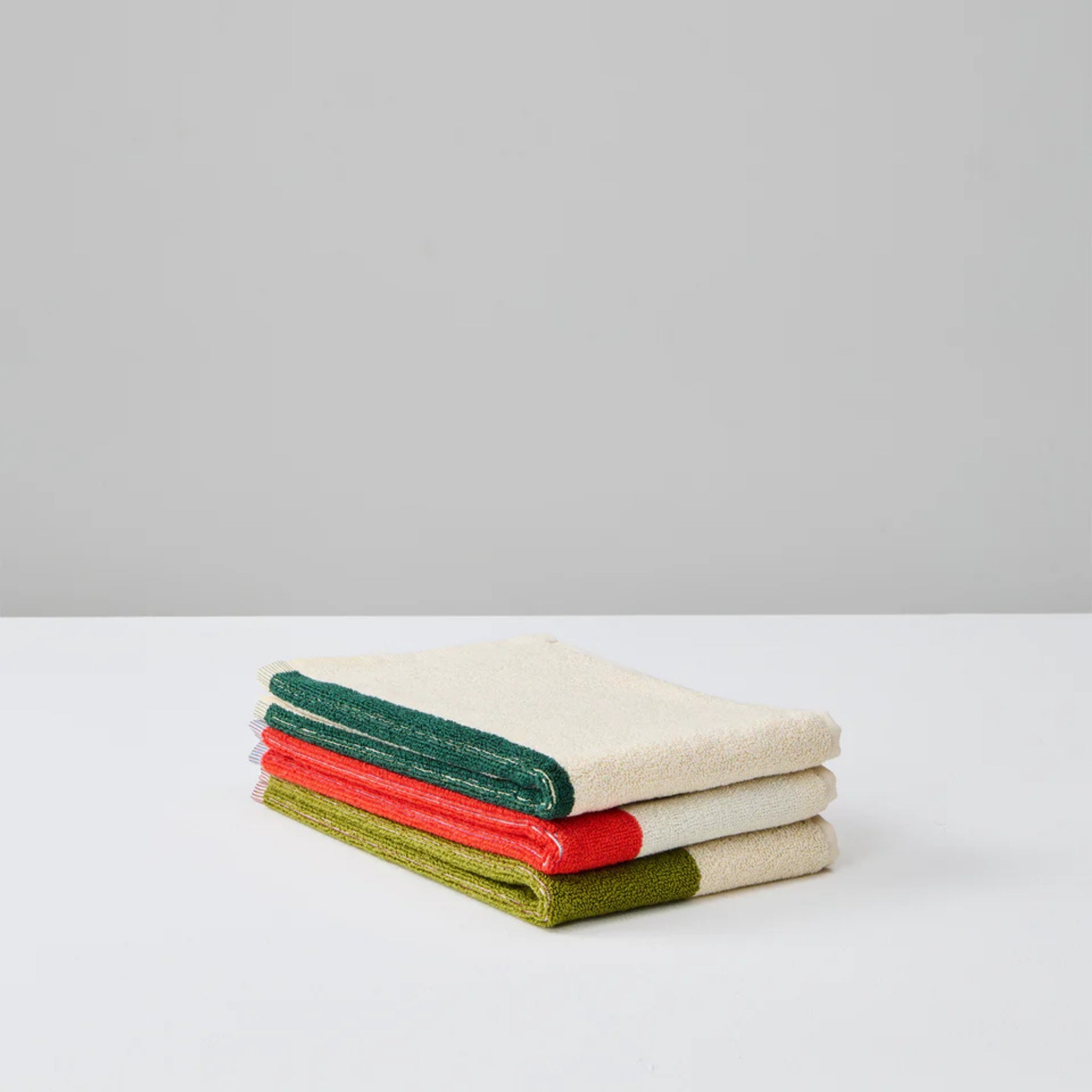 Een stapeltje van drie Suite 702 Gastendoekjes (Set D 30/50) gemaakt van biologisch katoen, met een beige hart en groene, rode en donkergroene randen, rustend op een wit oppervlak en effen grijze achtergrond.