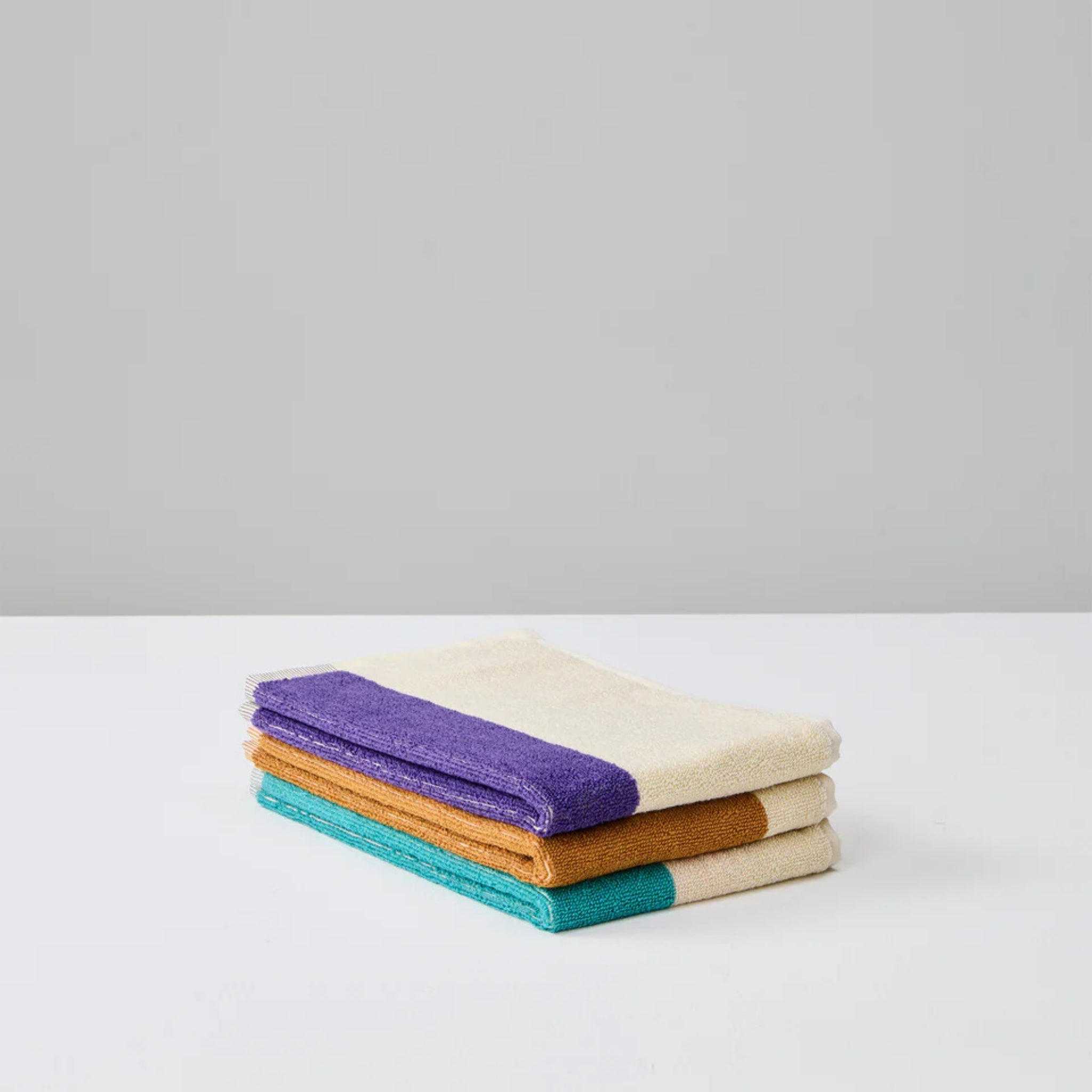 Drie crèmekleurige Suite 702 Gastendoekjes (Set C, 30/50, 3 st.) gemaakt van biologisch katoen zijn netjes gevouwen, elk met een opvallende paarse, oranje of groenblauwe streep op een wit oppervlak tegen een lichtgrijze achtergrond.