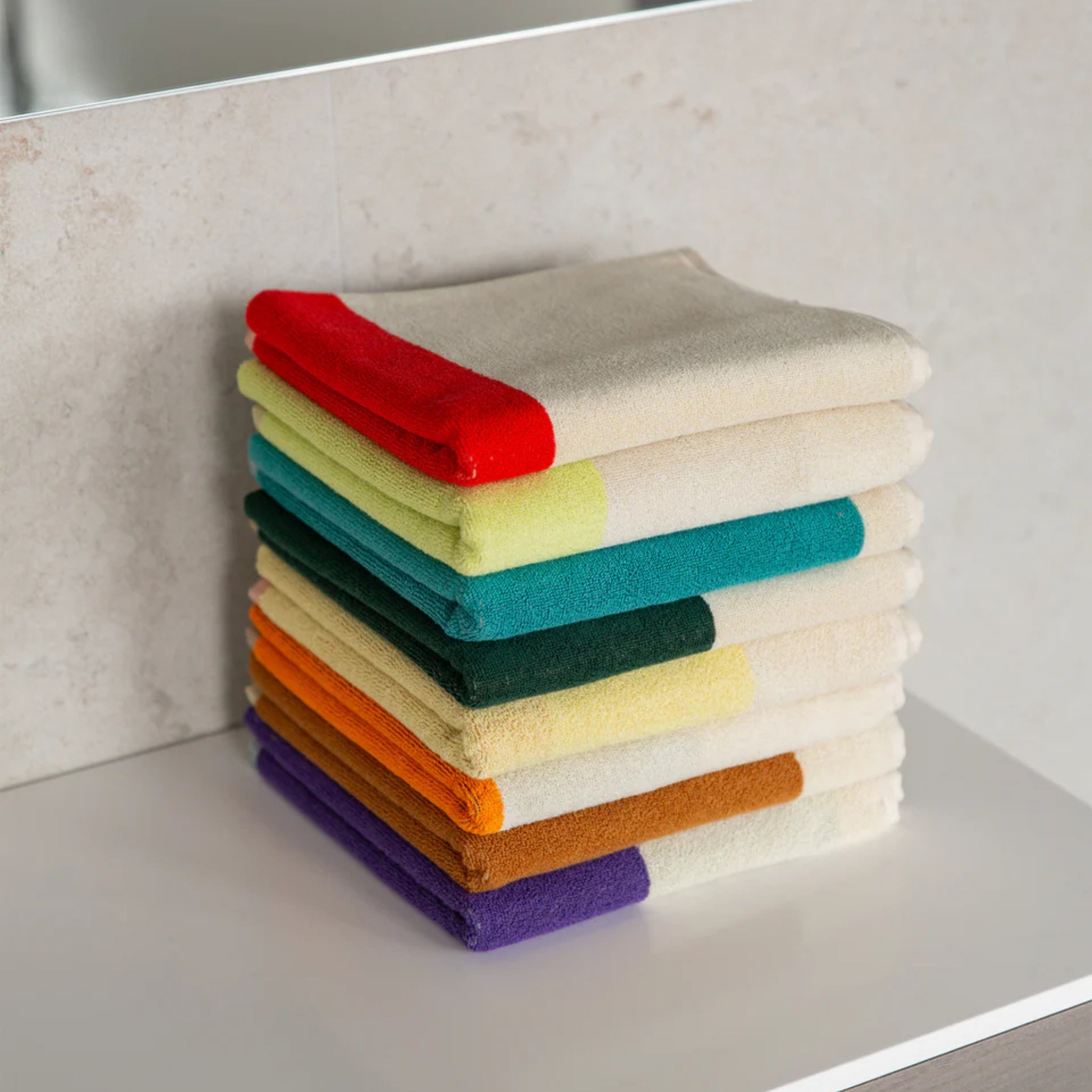 Een netjes gestapelde stapel Suite 702 Handdoek Rood Geel 50/100, elk met een levendige gekleurde streep, ligt op een wit oppervlak met lichte tegels en een spiegel op de achtergrond.