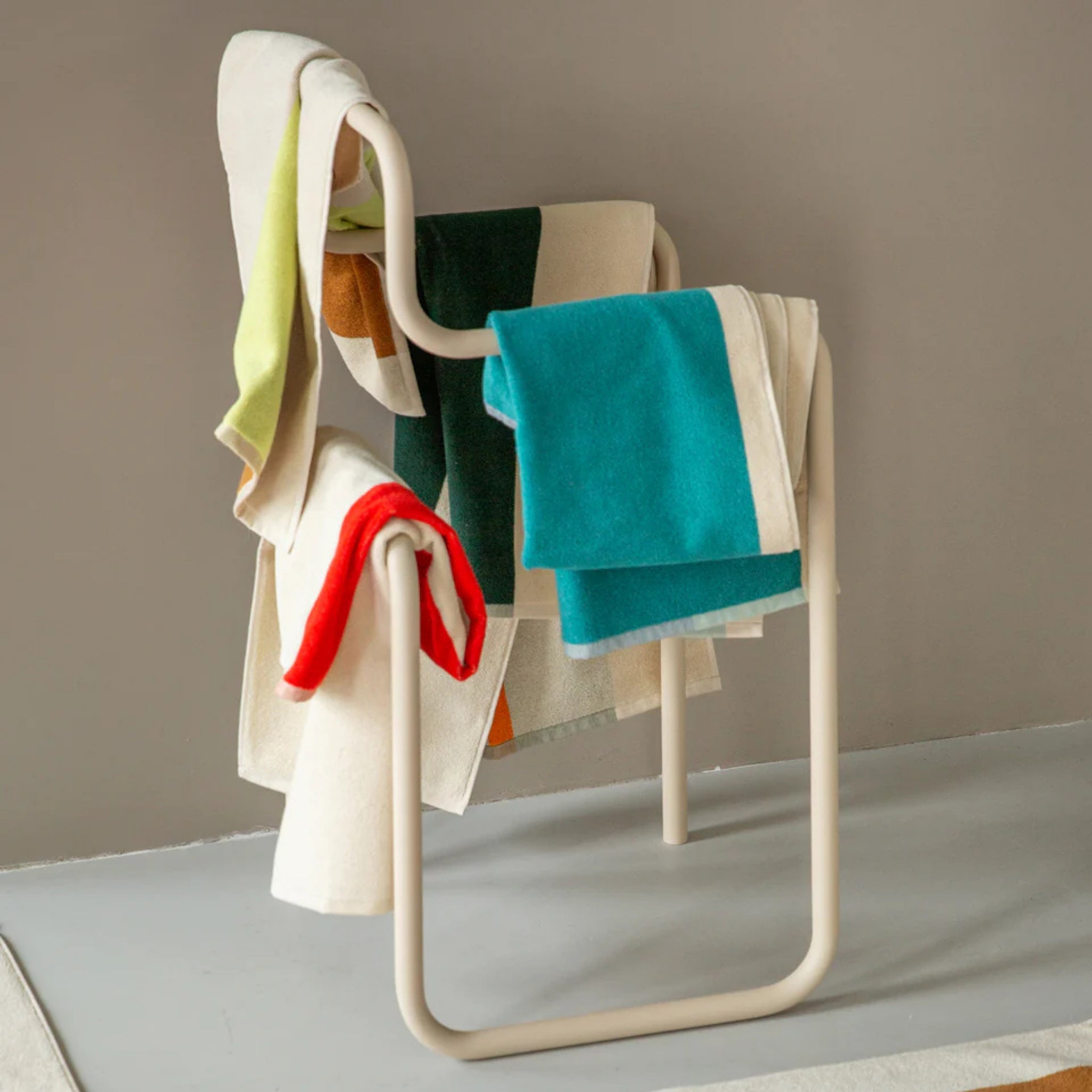 Een moderne witte metalen stoel gedrapeerd met kleurrijke handdoeken en dekens, waaronder de Suite 702 Handdoek Rood Geel 50/100 van Suite 702, steekt mooi af tegen een neutrale achtergrond.