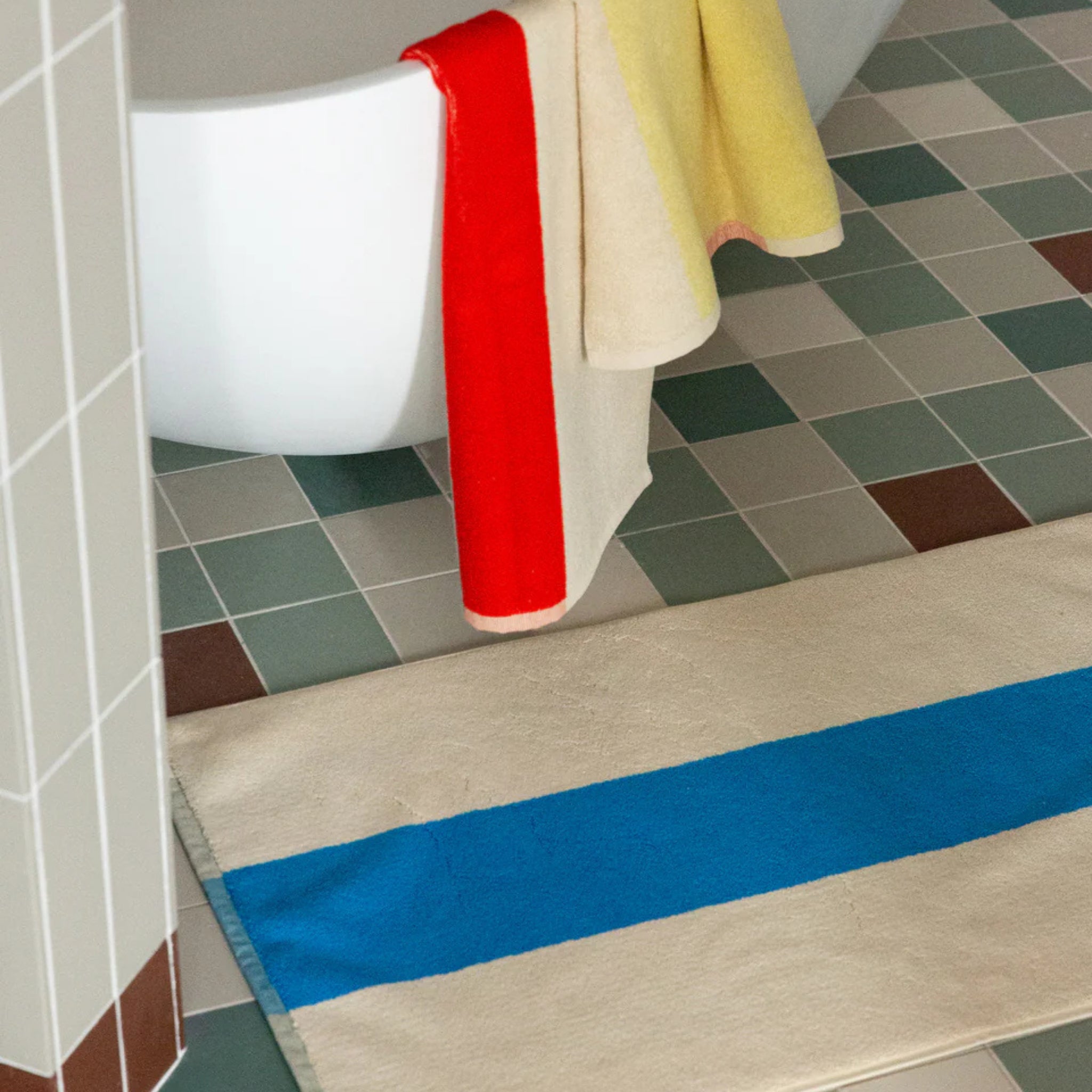 Een badkuip met een Suite 702 Handdoek Rood Geel 50/100 en een gele handdoek die over de rand hangt, naast een betegelde vloer en een crèmekleurige badmat met een felblauwe streep, allemaal uit Suite 702.
