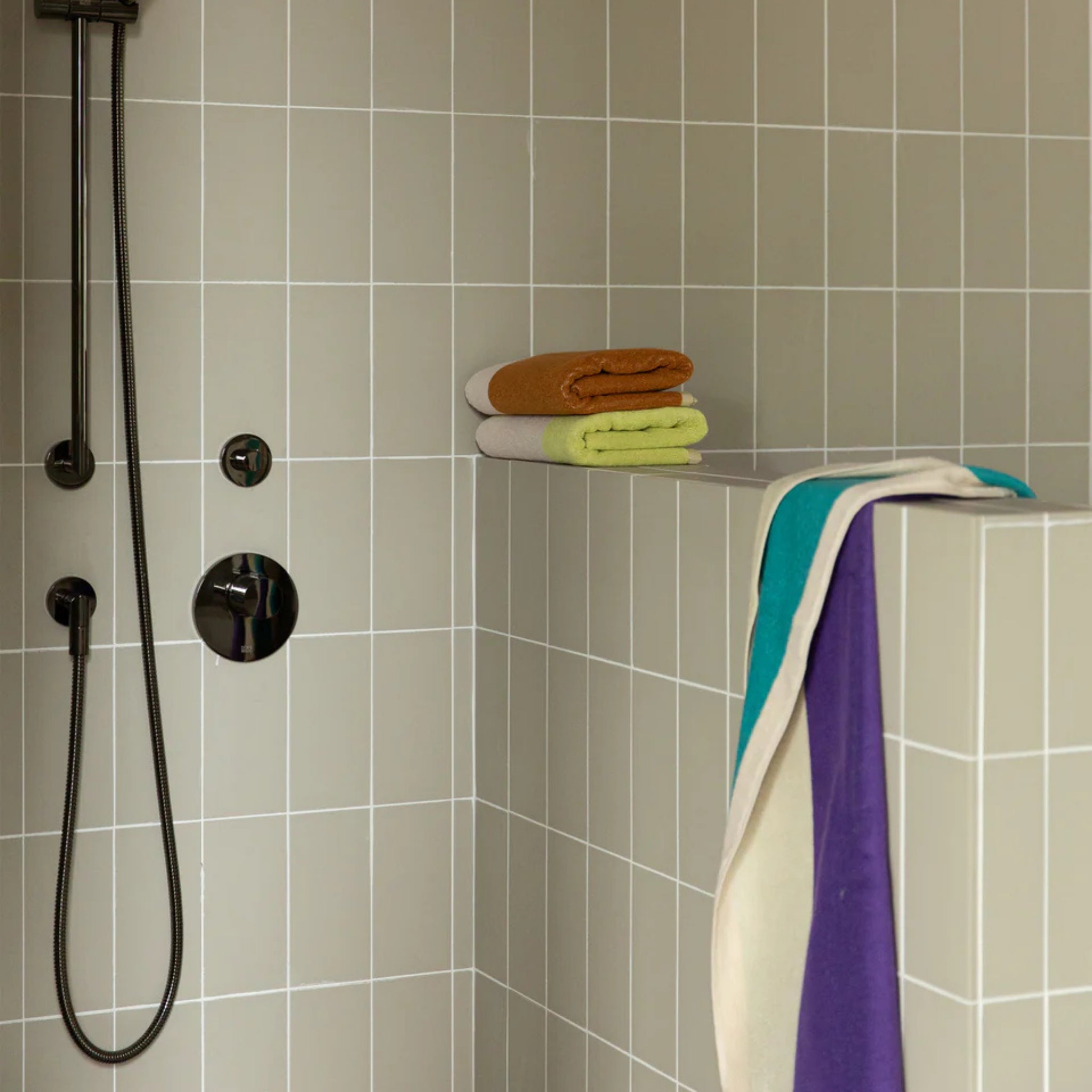 Een moderne douche met lichtgrijze vierkante tegels, zwarte douchekop en handgreep, en een halve muur bedekt met gevouwen biokatoenen handdoeken; een Suite 702 Handdoek Lime Toffee 50/100 van Suite 702 hangt over de muur.