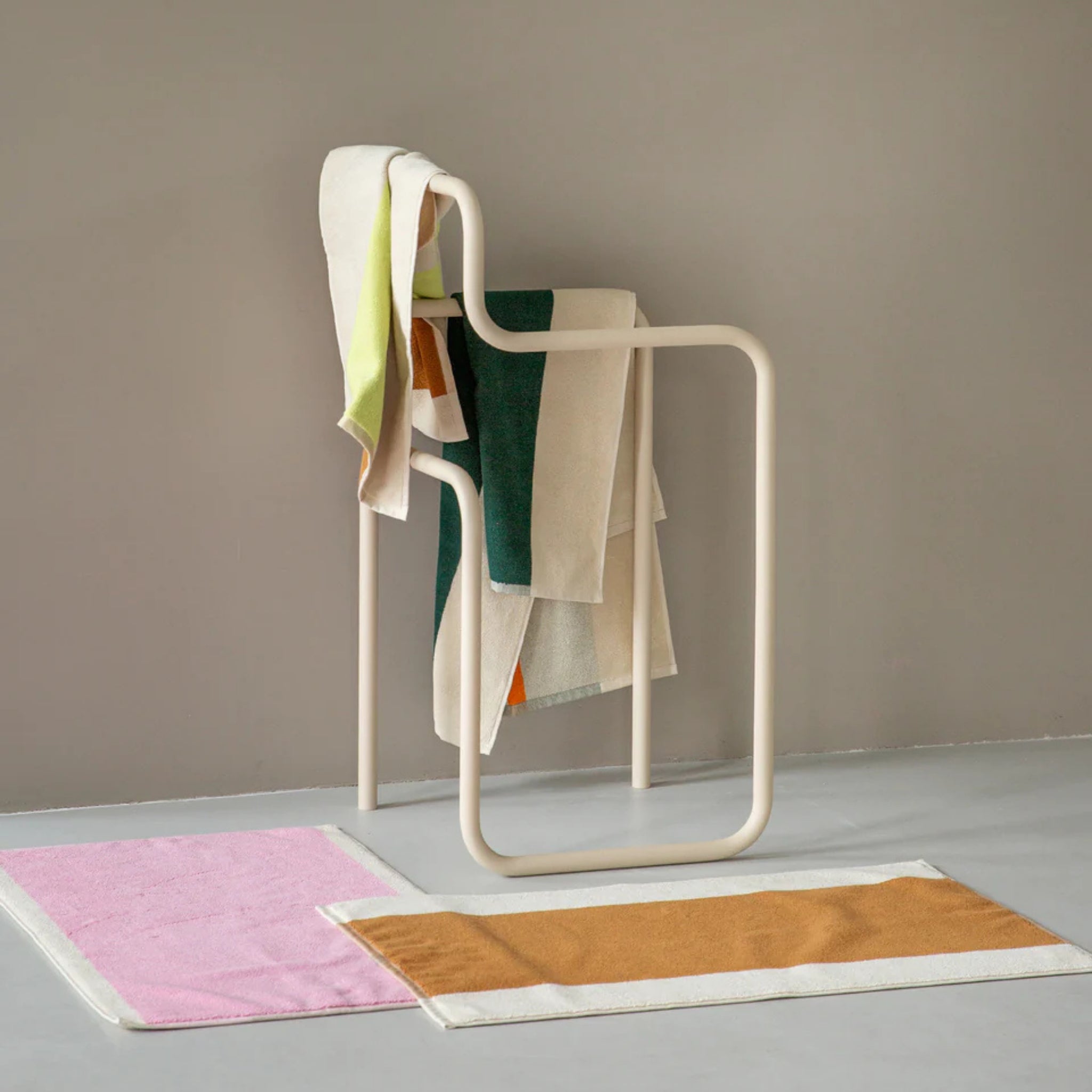 Een modern, sculpturaal handdoekenrek toont verschillende kleurrijke handdoeken, waaronder de Suite 702 Handdoek Donkergroen Oranje 50/100. Twee rechthoekige badmatten - een roze en een bruine met witte randen - liggen op een lichtgrijze vloer tegen een beige muur.