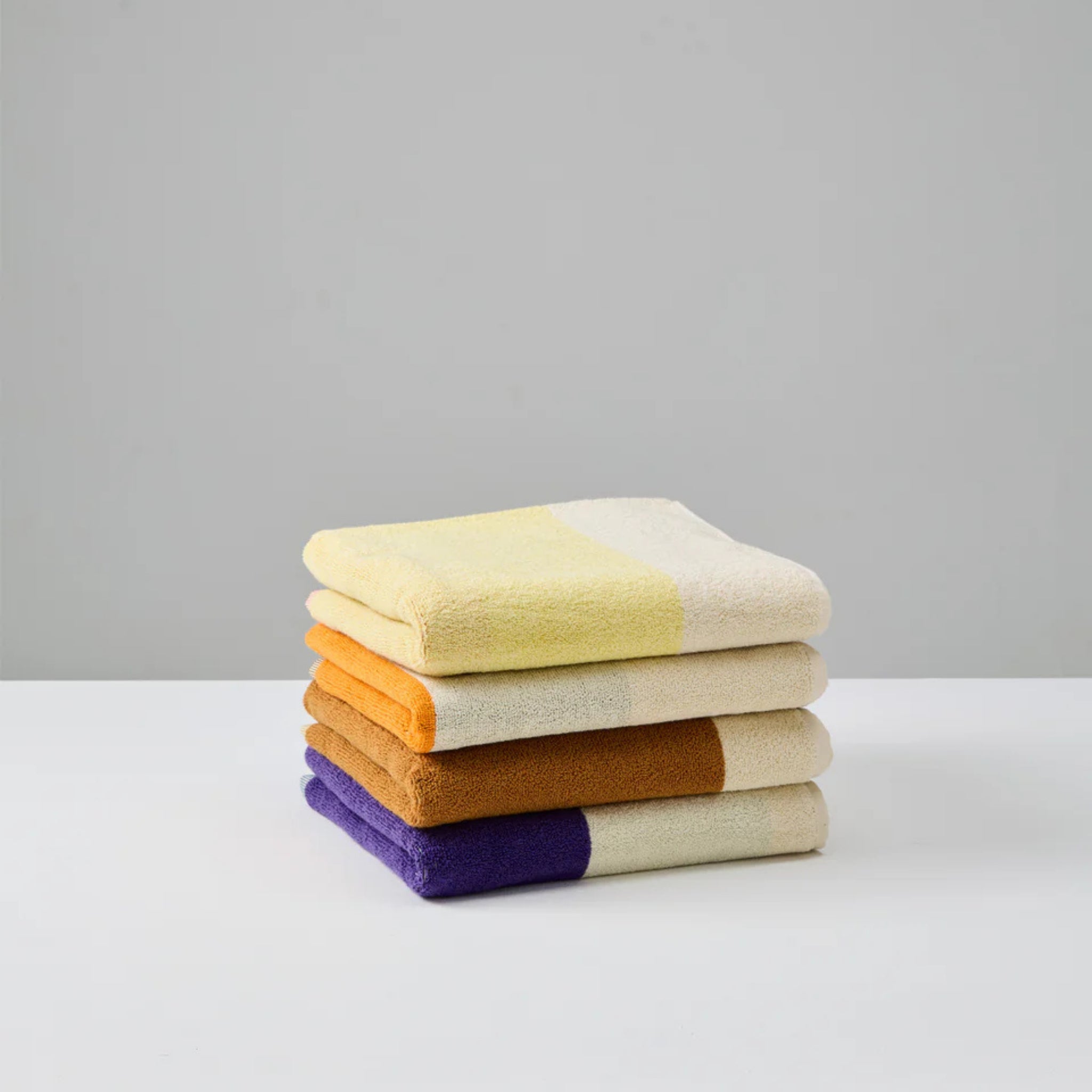 Een keurig gestapelde stapel van vier Suite 702 Handdoeken Rood Geel 50/100, met op Karel Martens geïnspireerde gele, oranje, bruine en paarse strepen, ligt op een wit oppervlak met een effen lichtgrijze achtergrond.