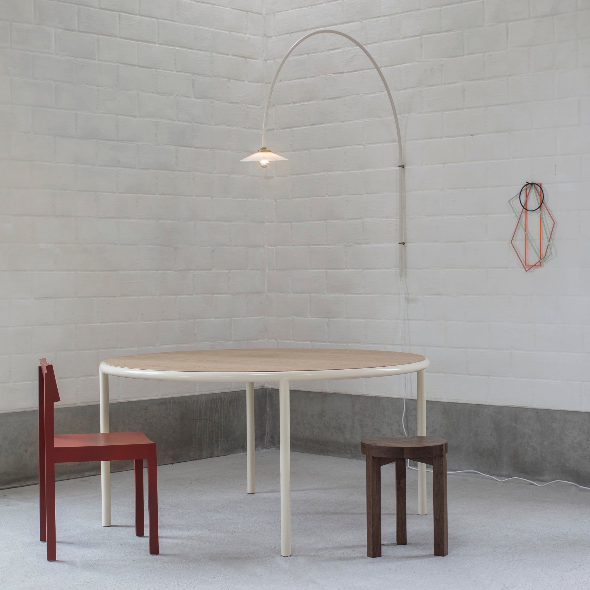 Een minimalistische kamer met een ronde houten tafel, rode stoel en bruine kruk toont Valerie Objects Muller van Severen Onderzetter Multicolor 4 st. op een witte bakstenen muur, verlicht door een wandlamp boven een grijze vloer en lagere muren.