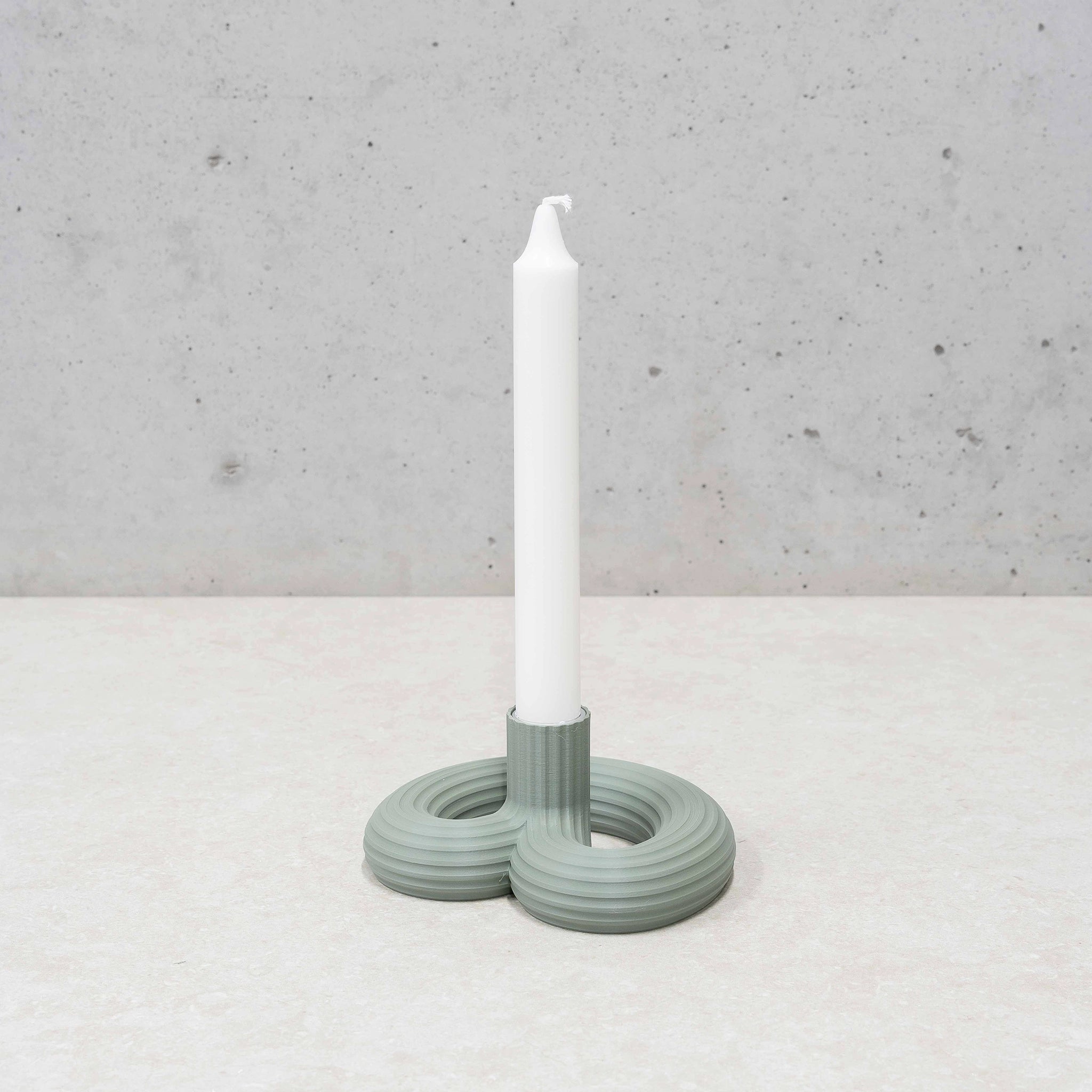 Een witte conische kaars staat in de Wavy Kandelaar Muntgroen van Wavy Design Studio, een moderne lichtgroene kandelaar met drie onderling verbonden ringen, op een licht oppervlak tegen een betonnen muur.