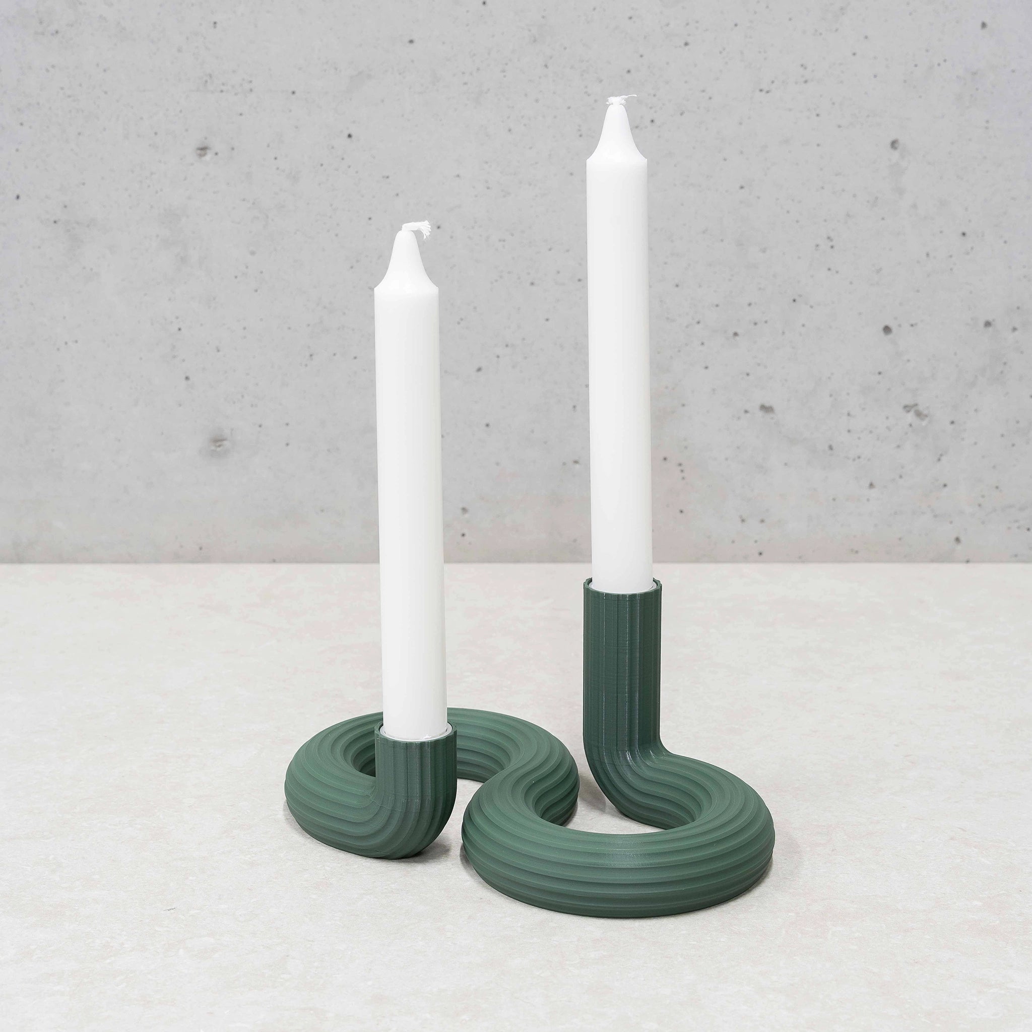 Twee witte kaarsen in de Wavy Dubbele Kandelaar Highland Groen van Wavy Design Studio voegen een vleugje duurzaam interieur toe aan een licht oppervlak en steken af tegen een grijze, gespikkelde achtergrond.