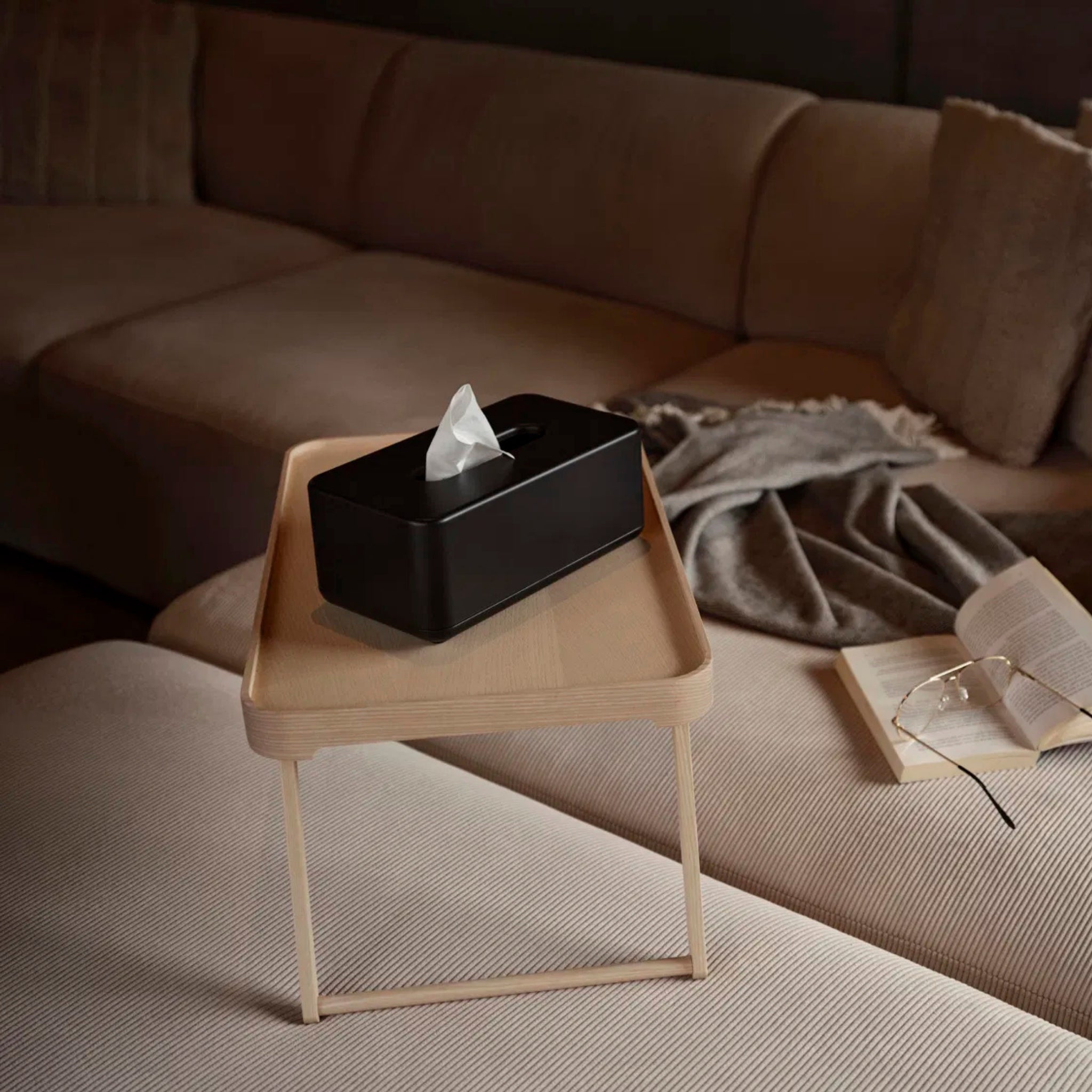 Een Zone Denmark Ume Tissuebox Zwart rust op een klein houten dienbladtafeltje voor een beige sofa met een grijs kleed, een open boek en een bril, en creëert een gezellige sfeer met zachte verlichting.
