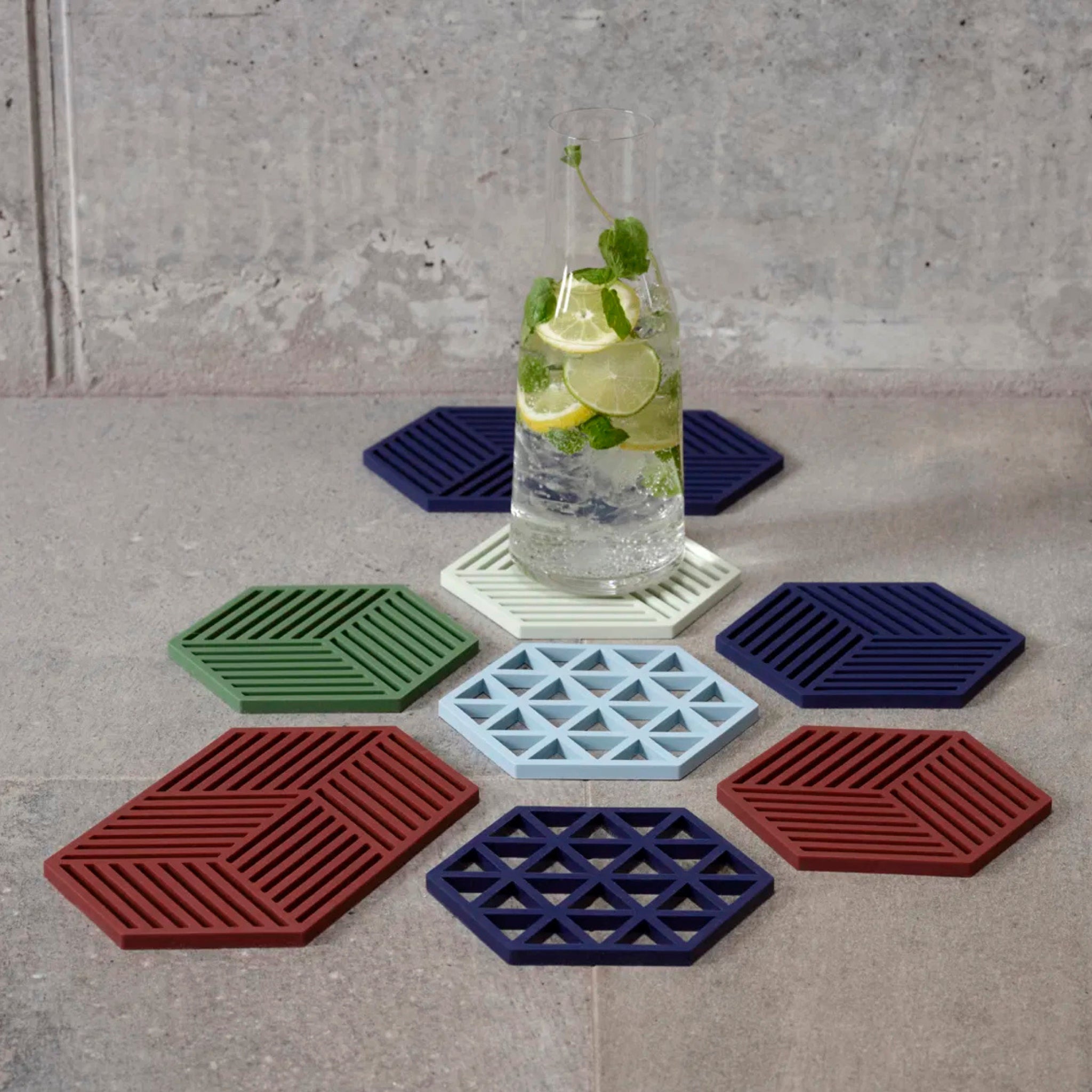Een glazen fles met citroen, limoen en munt wordt geplaatst op de Zone Denmark Triangles Trivet Indigo van Zone Denmark - deze moderne, hittebestendige siliconen onderzetters in indigo zijn in een cirkel geplaatst op een grijs betegeld oppervlak.