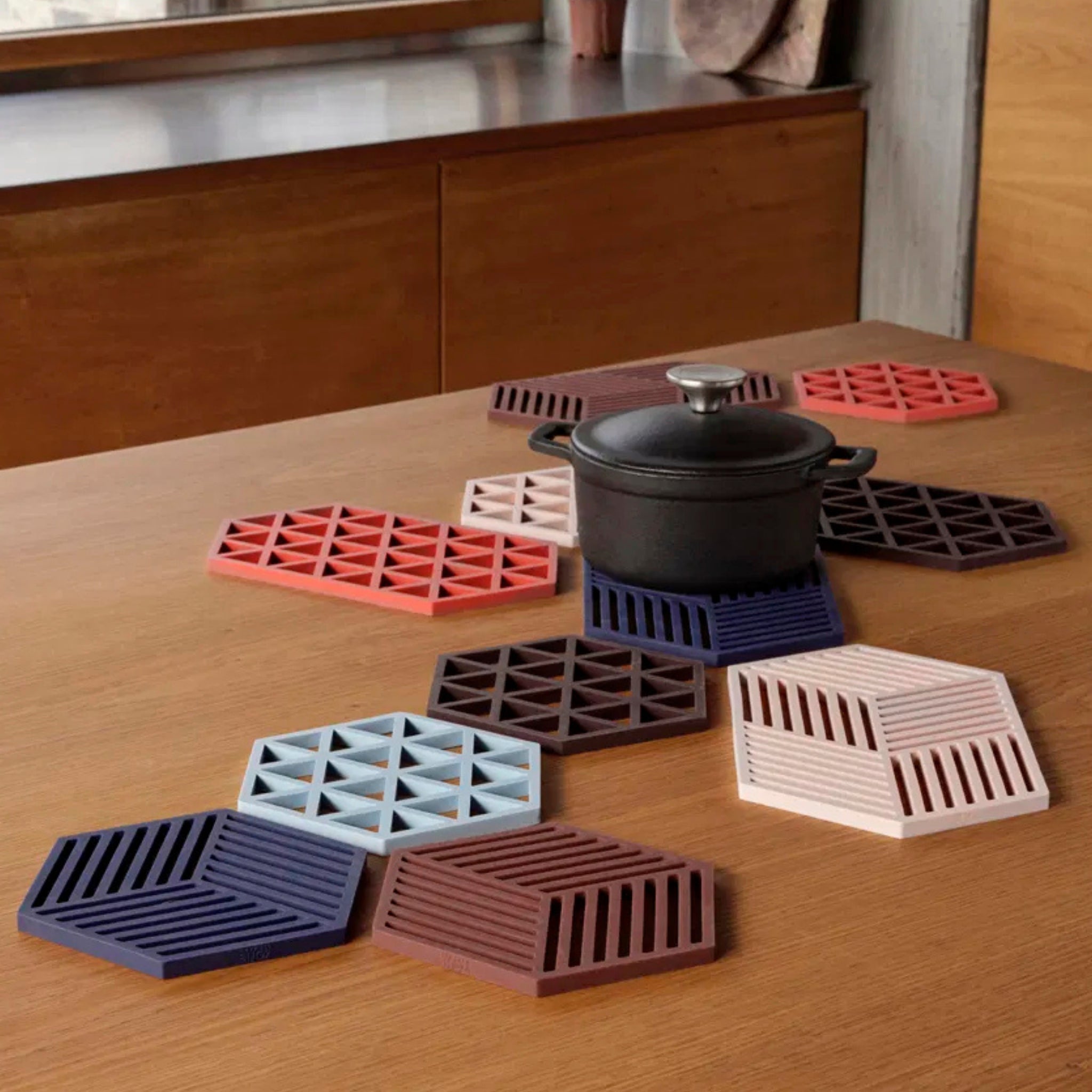 Een zwarte pan met deksel staat op een houten tafel, rustend op Zone Denmark Triangles Trivet Blauw siliconen onderzetters in verschillende kleuren en geometrische patronen. Op de achtergrond staan een houten kast en een raam.