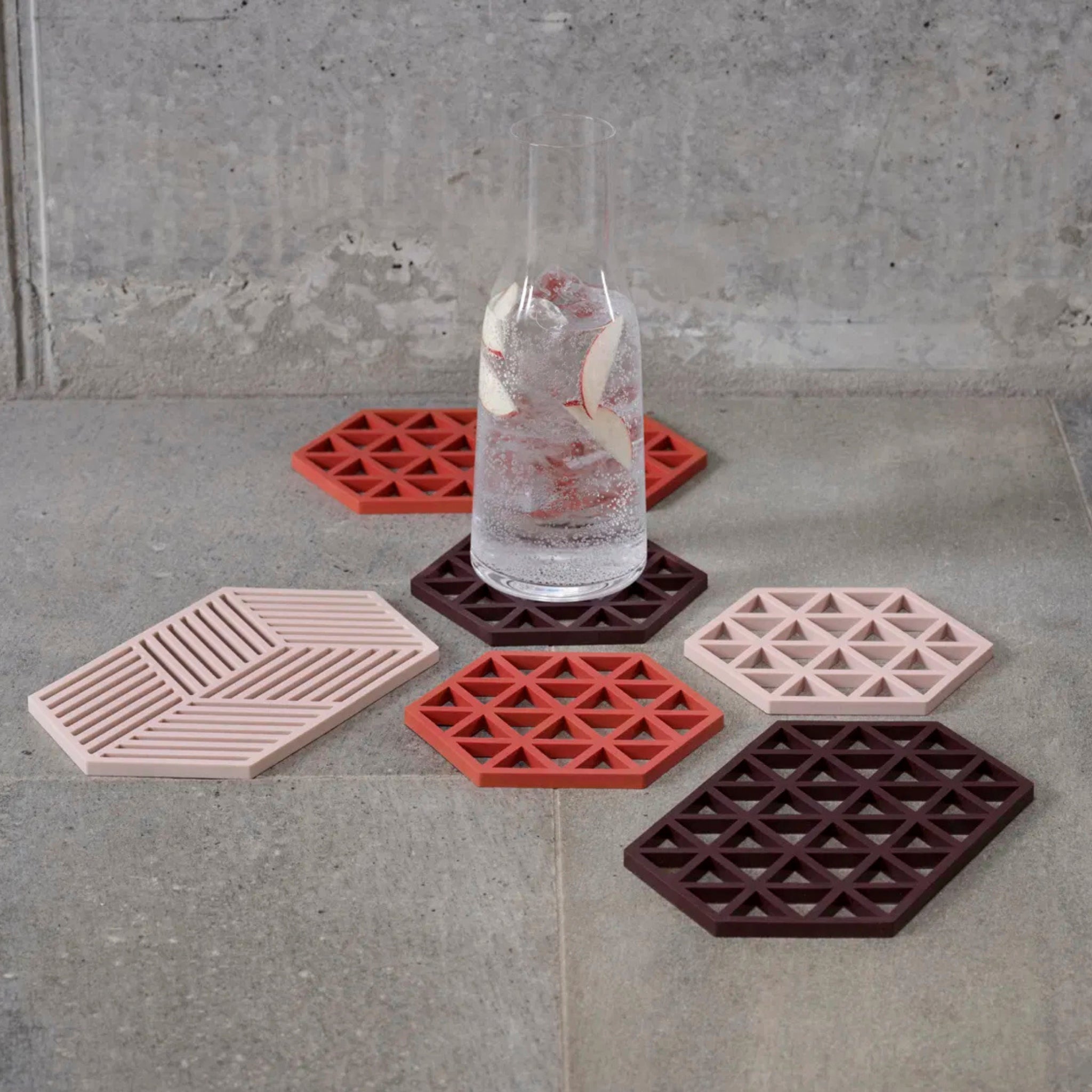 Een hoog glas bruisend water met ijs en fruit staat op een grijze tegel, omringd door zes hittebestendige Zone Denmark Triangles Trivet Kalamata siliconen onderzetters in rood-, roze- en bordeauxtinten.