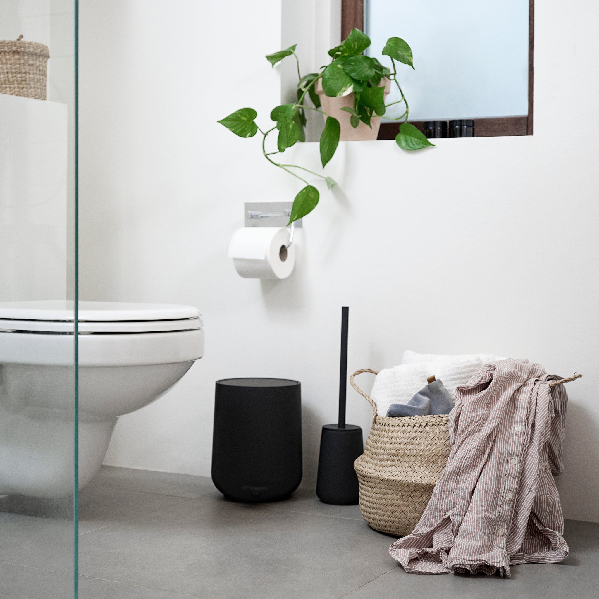 Een moderne badkamer met een wit toilet, wandclosethouder, Zone Denmark Nova One Pedaalemmer Zwart 3 l en toiletborstel, een plant bij het raam, plus gevlochten manden met handdoeken en een gestreepte doek op de grijze vloer.