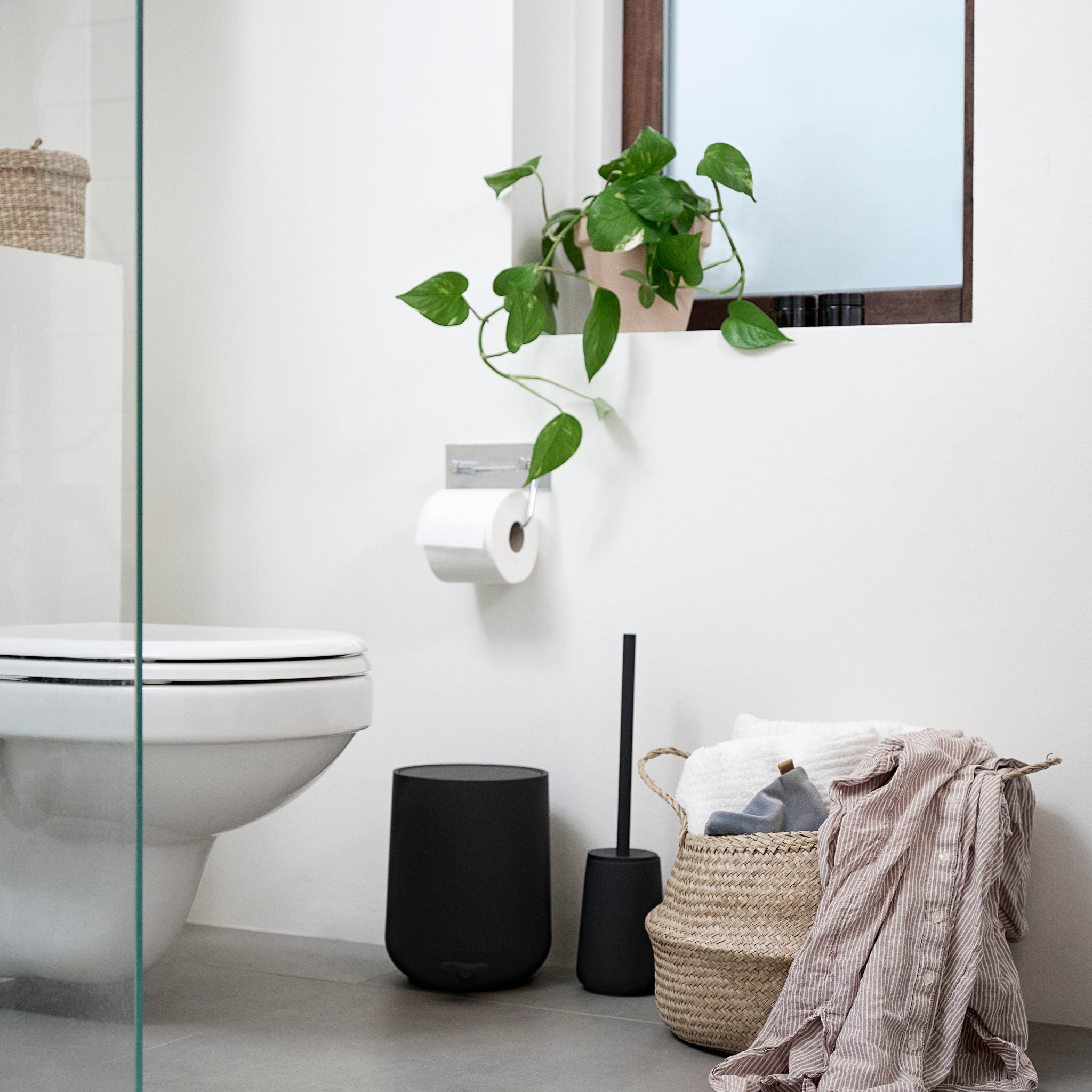 Moderne badkamer met een wit toilet, zwarte prullenbak en de Zone Denmark Nova One Toiletborstel Zwart als onmisbaar accessoire. Handdoeken en een gestreepte badjas vullen de mand; een plant in pot voegt frisheid toe boven het toiletpapier.