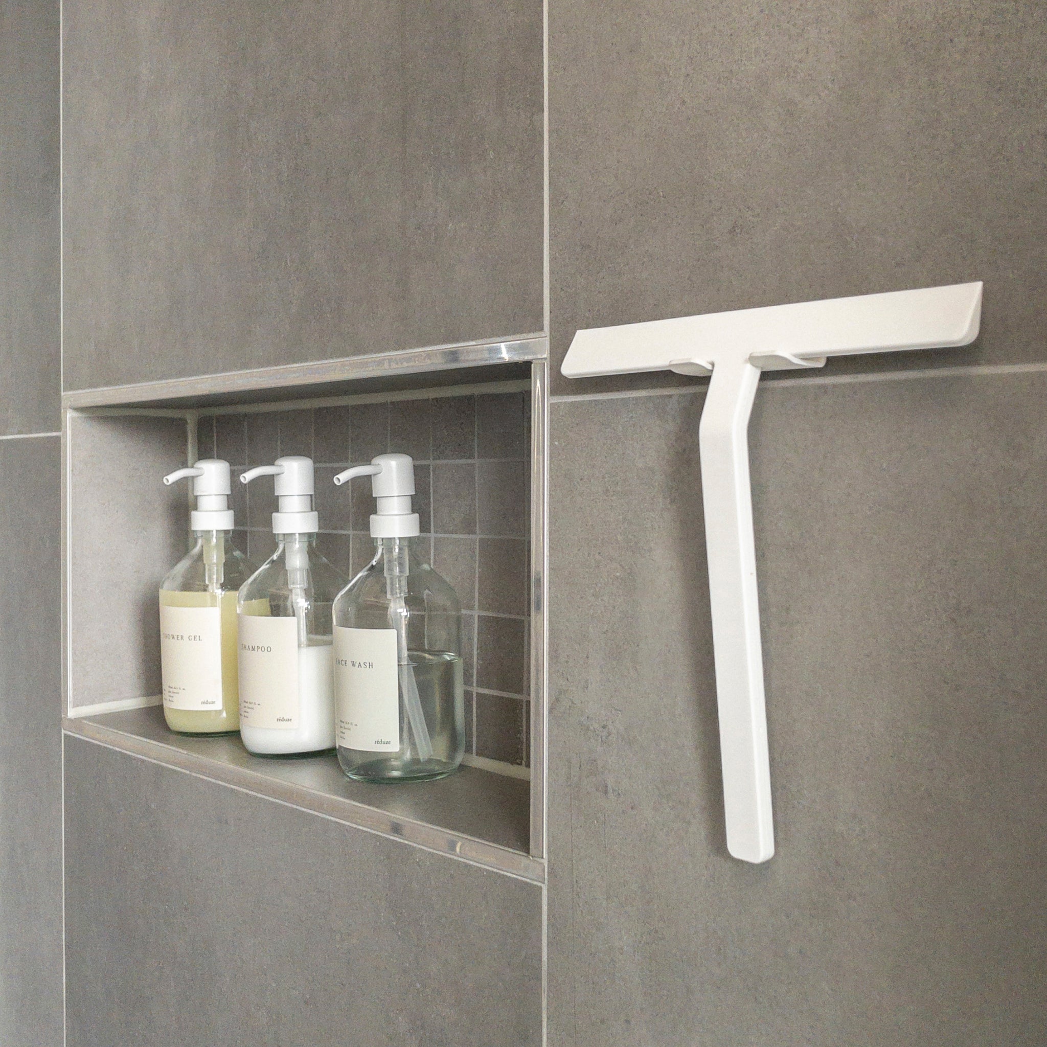 Een douchenis met drie heldere pompflessen voor zeep, shampoo en conditioner staat naast de stijlvolle Zone Denmark Rim Douchewisser met Houder Wit S-praktische badkameraccessoires van Zone Denmark op een grijs betegelde muur.