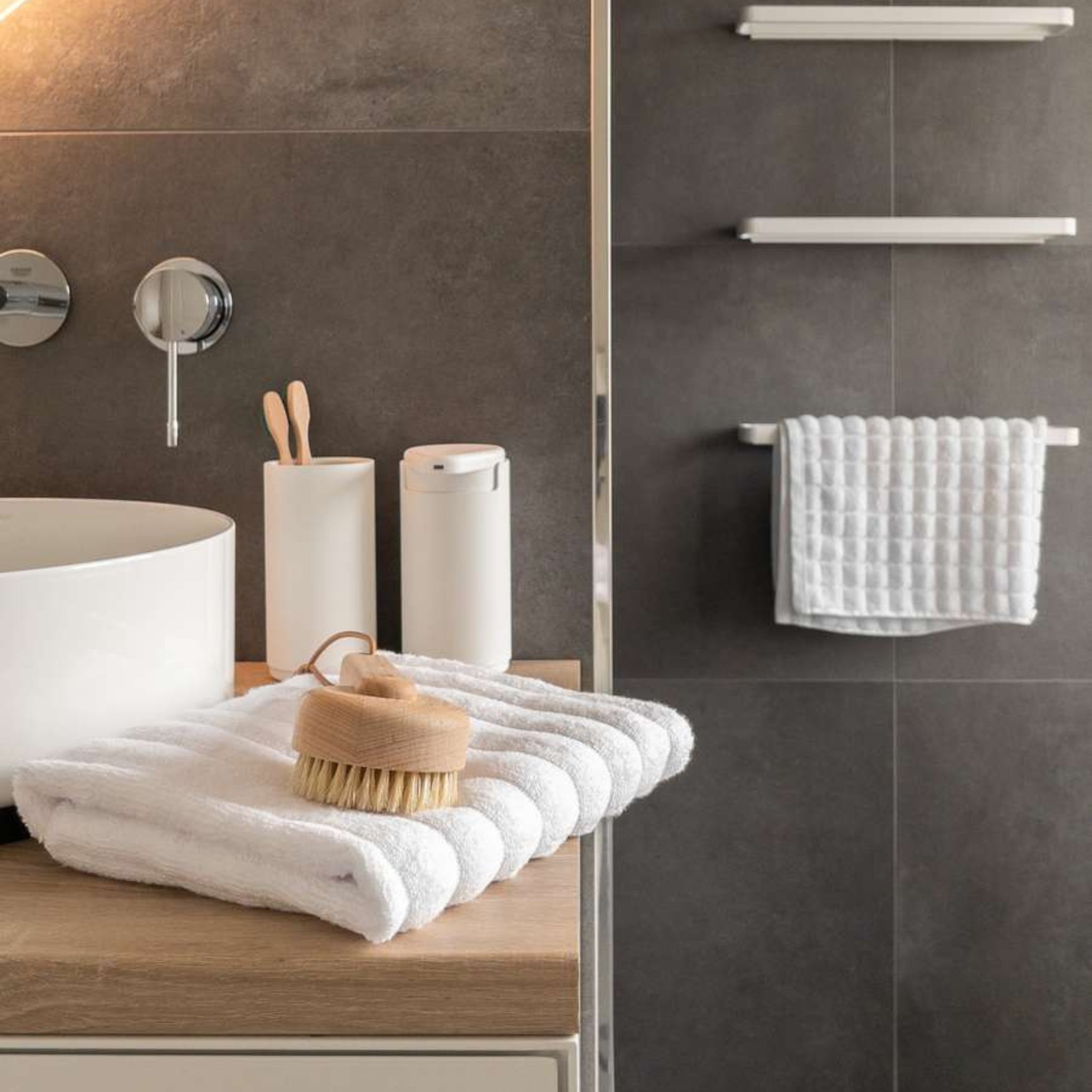 Moderne badkamer met een witte wastafel, badkamer organizer en zeepdispenser op een houten aanrecht. Opgevouwen handdoeken en een badborstel liggen op de voorgrond; een handdoek hangt bij de Zone Denmark Rim Tandenborstelhouder Wit.