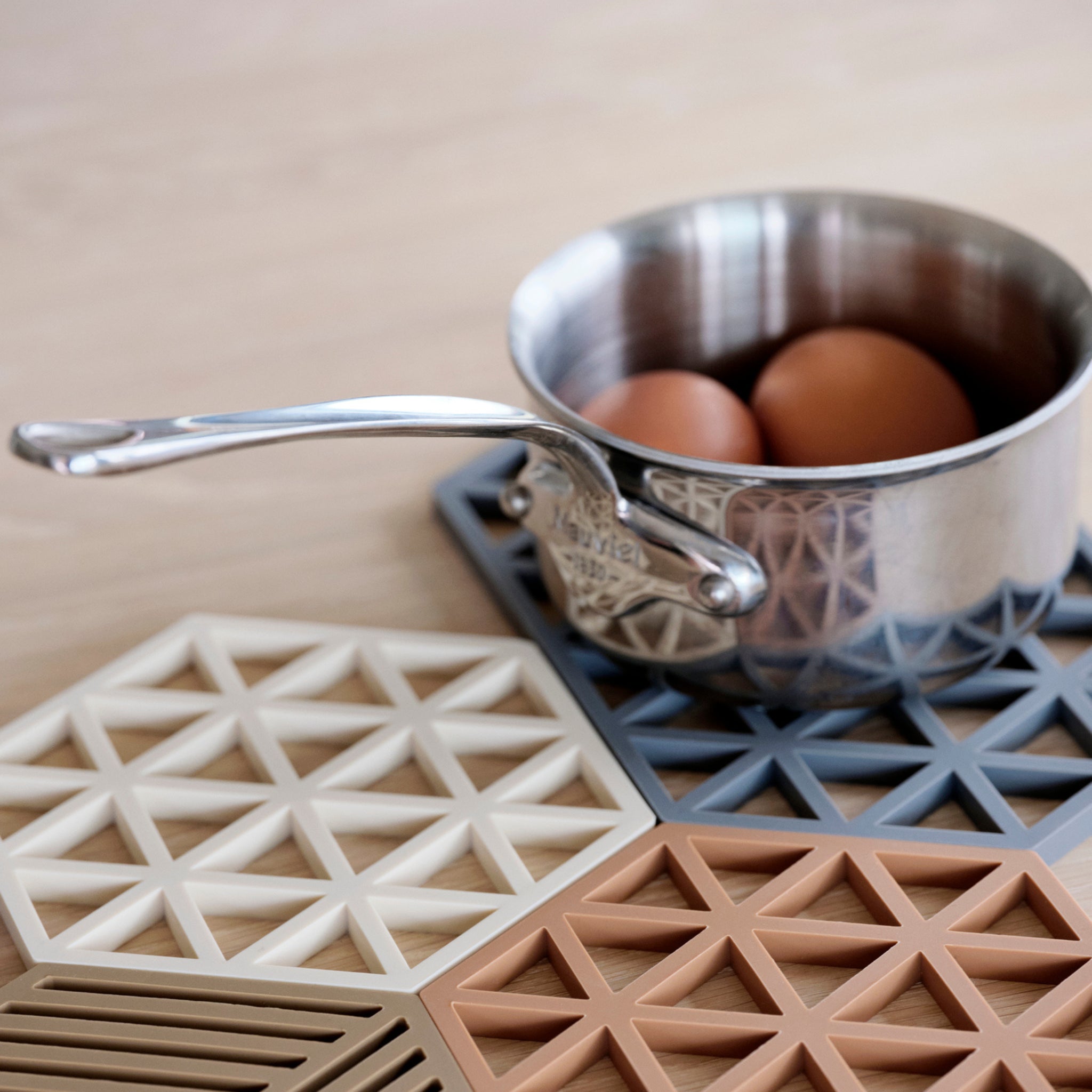 Een roestvrijstalen steelpan met twee bruine eieren staat op een houten oppervlak op een Zone Denmark Triangles Trivet Denim, gemaakt door Zone Denmark.