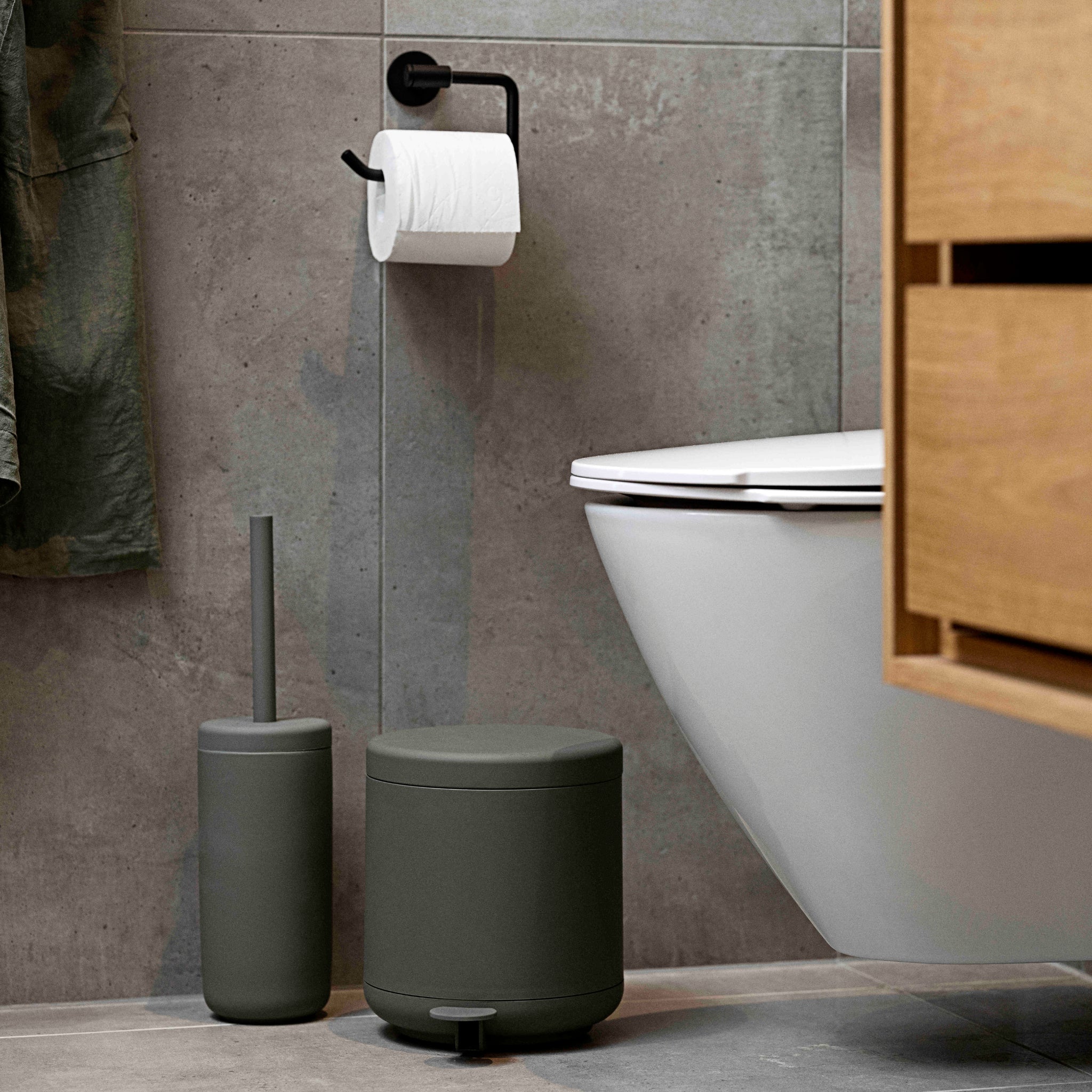 Een moderne badkamer met een wandcloset, grijze tegelwanden, een zwarte toiletrolhouder en de Zone Denmark Ume pedaalemmer in olijfgroen (4 L). Een bijpassende grijze toiletborstel uit de Ume-serie en een houten kastje zijn ook aanwezig.