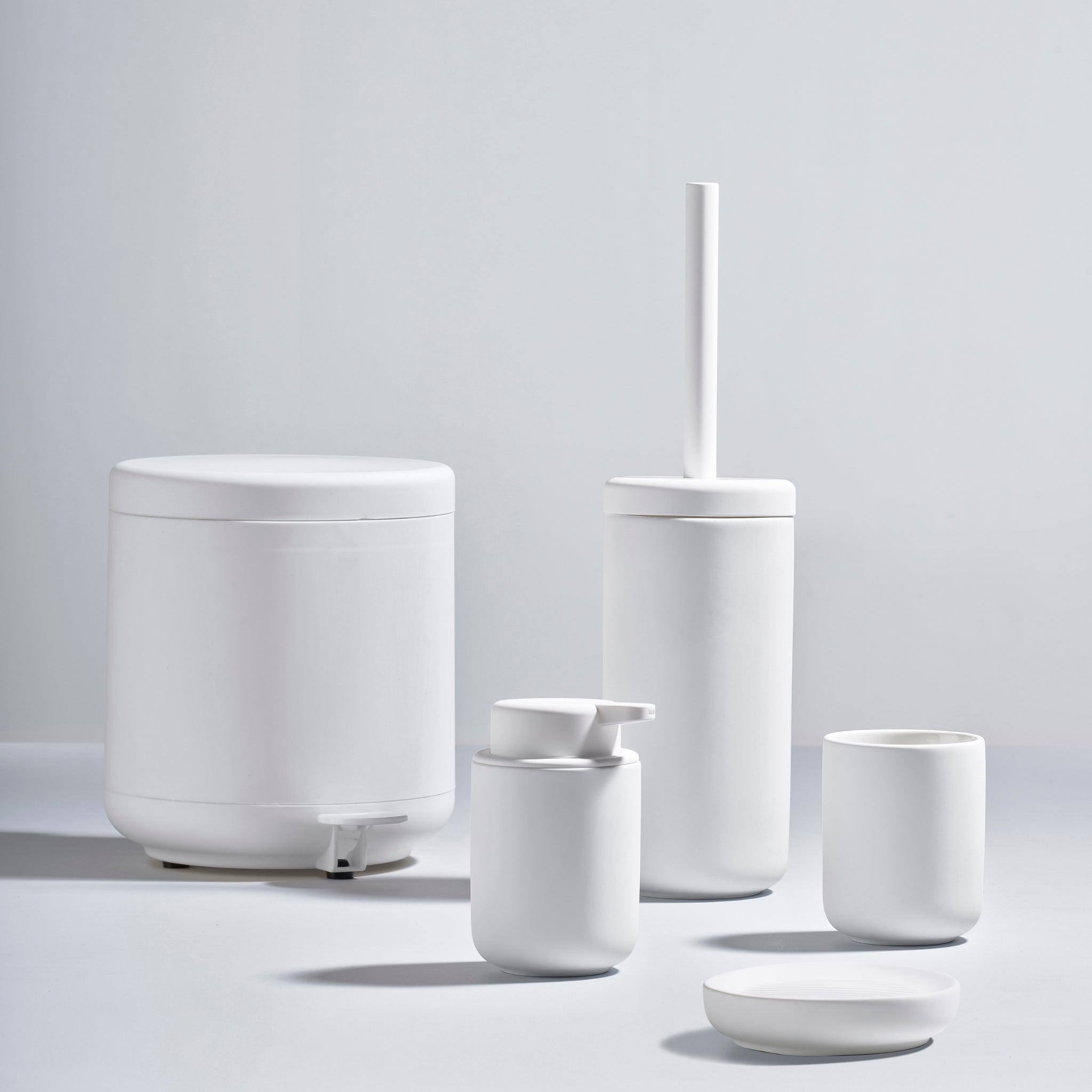 Minimalistische witte badkamerset met Zone Denmark Ume Pedaalemmer Wit 4 l, toiletborstelgarnituur, zeepdispenser, beker en zeepbakje - allemaal in een gladde matte afwerking tegen een lichtgrijze achtergrond.