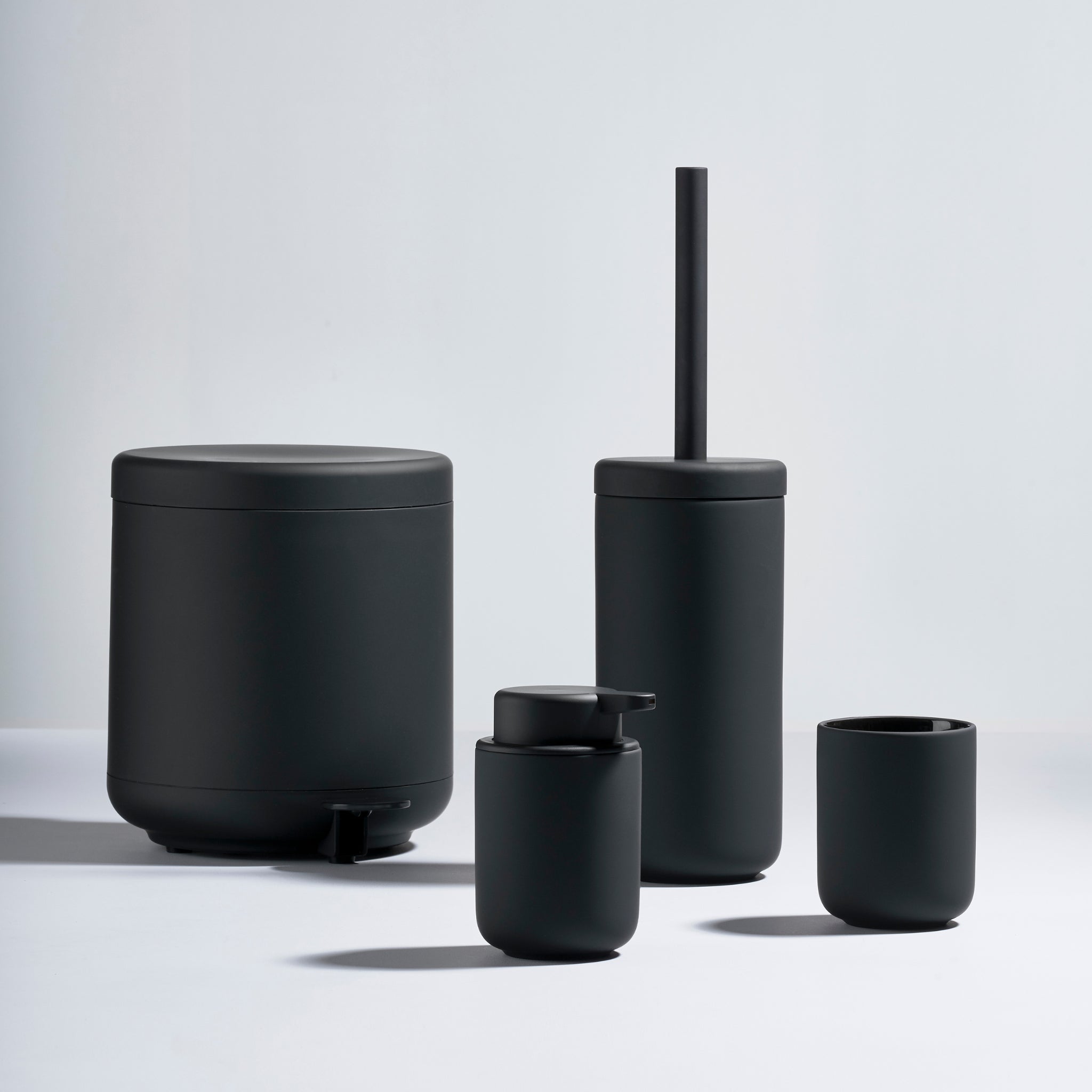 Een set van vier matzwarte badkameraccessoires van Zone Denmark, waaronder de Ume Pedaalemmer Zwart 4 l, toiletborstelhouder met borstel, zeepdispenser en beker, gerangschikt op een lichtgrijs oppervlak met een effen achtergrond.