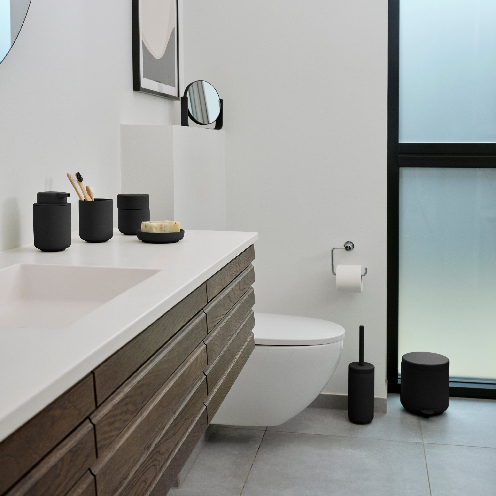 Een moderne badkamer met een witte wastafel, houten badmeubel, zwarte Zone Denmark Ume accessoires waaronder de Ume Pedaalemmer Zwart 4L, wandcloset, toiletrol en een matglazen raam dat natuurlijk licht binnenlaat.
