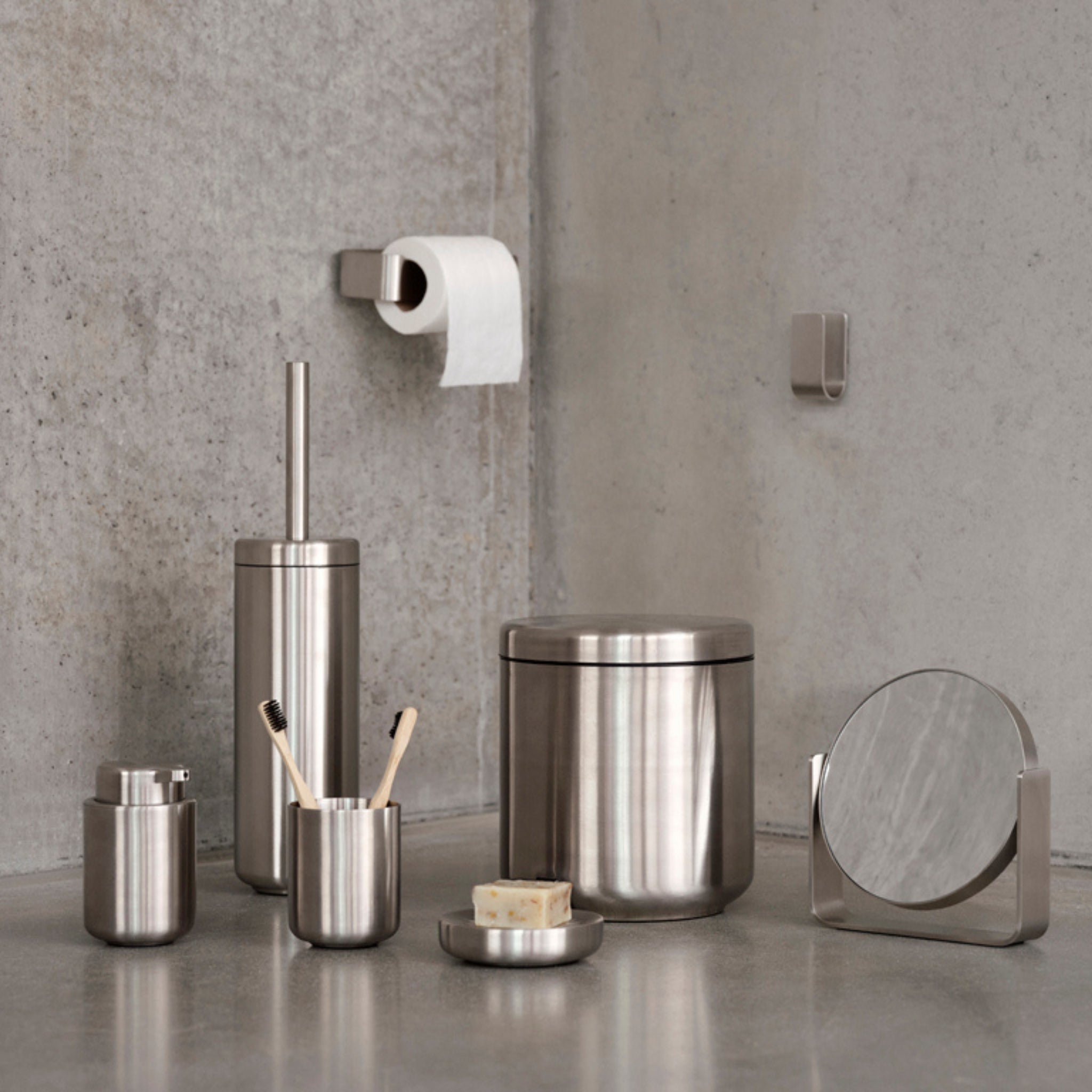 De Zone Denmark Ume Tandenborstelhouder Staal en andere roestvrijstalen badkameraccessoires van Zone Denmark staan uitgestald op een grijs aanrechtblad, met daarboven een wc-papierrol.