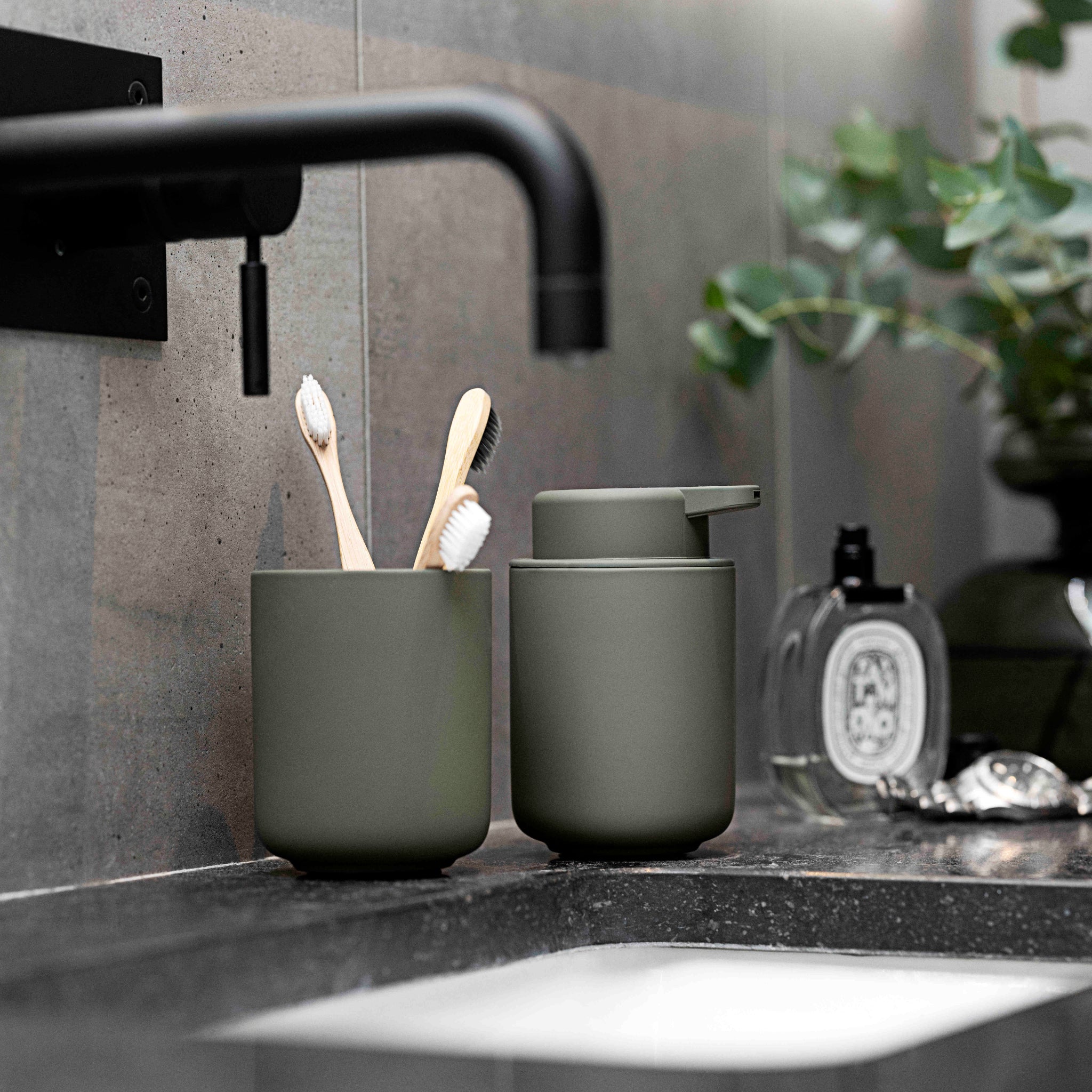 Twee houten tandenborstels in een matte olijfgroene Zone Denmark Ume Tandenborstelhouder zitten naast een bijpassende zeepdispenser op een moderne zwarte wastafel met zwarte kraan, plant en parfumfles, allemaal van de Ume badkamerserie.